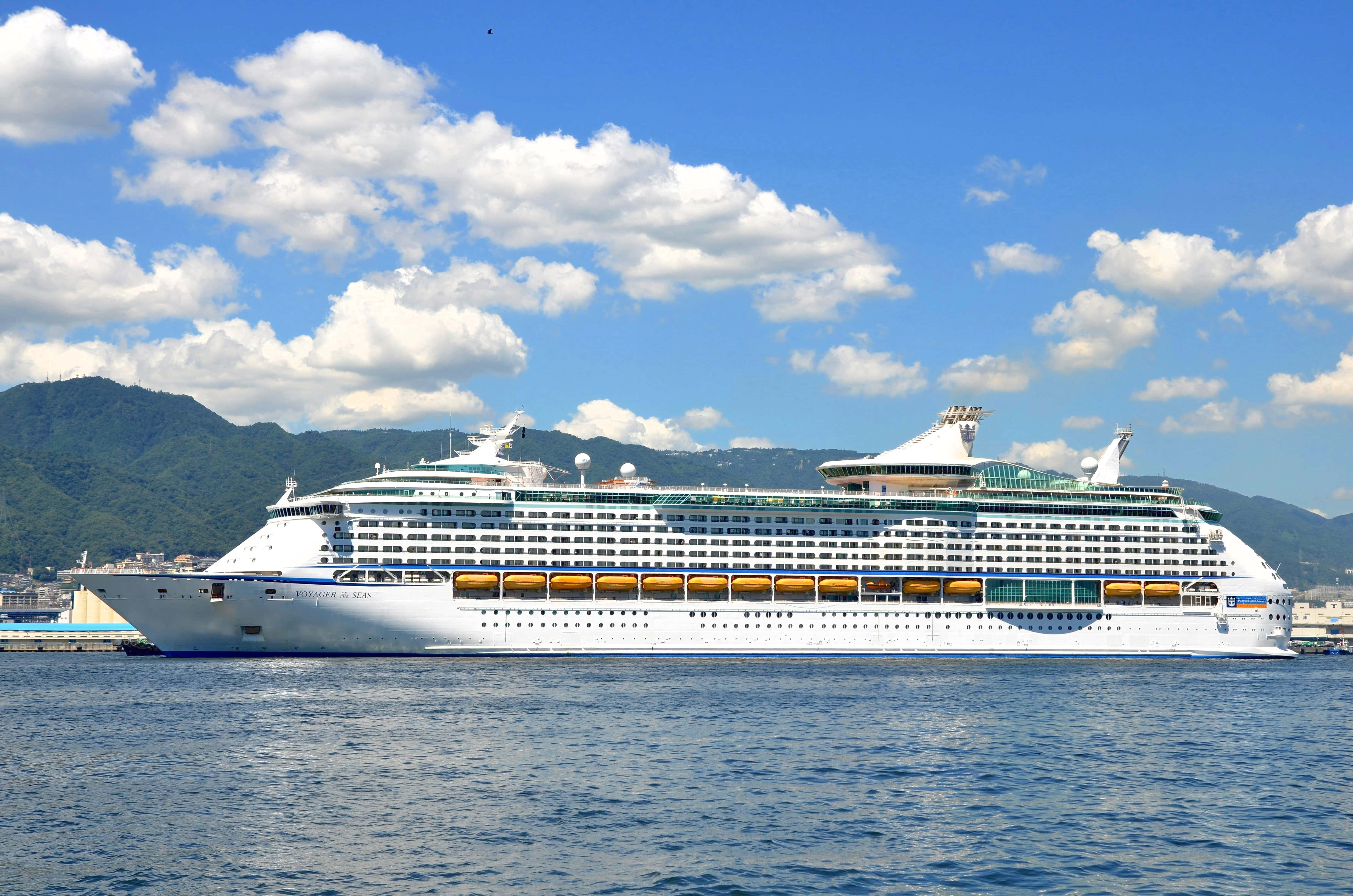 Cruise Ship 桌布 壁紙 — 一艘巨大的白色郵輪帶有藍色飾邊，在深藍色海面上平靜地漂浮，天空明亮，有蓬鬆的白雲。