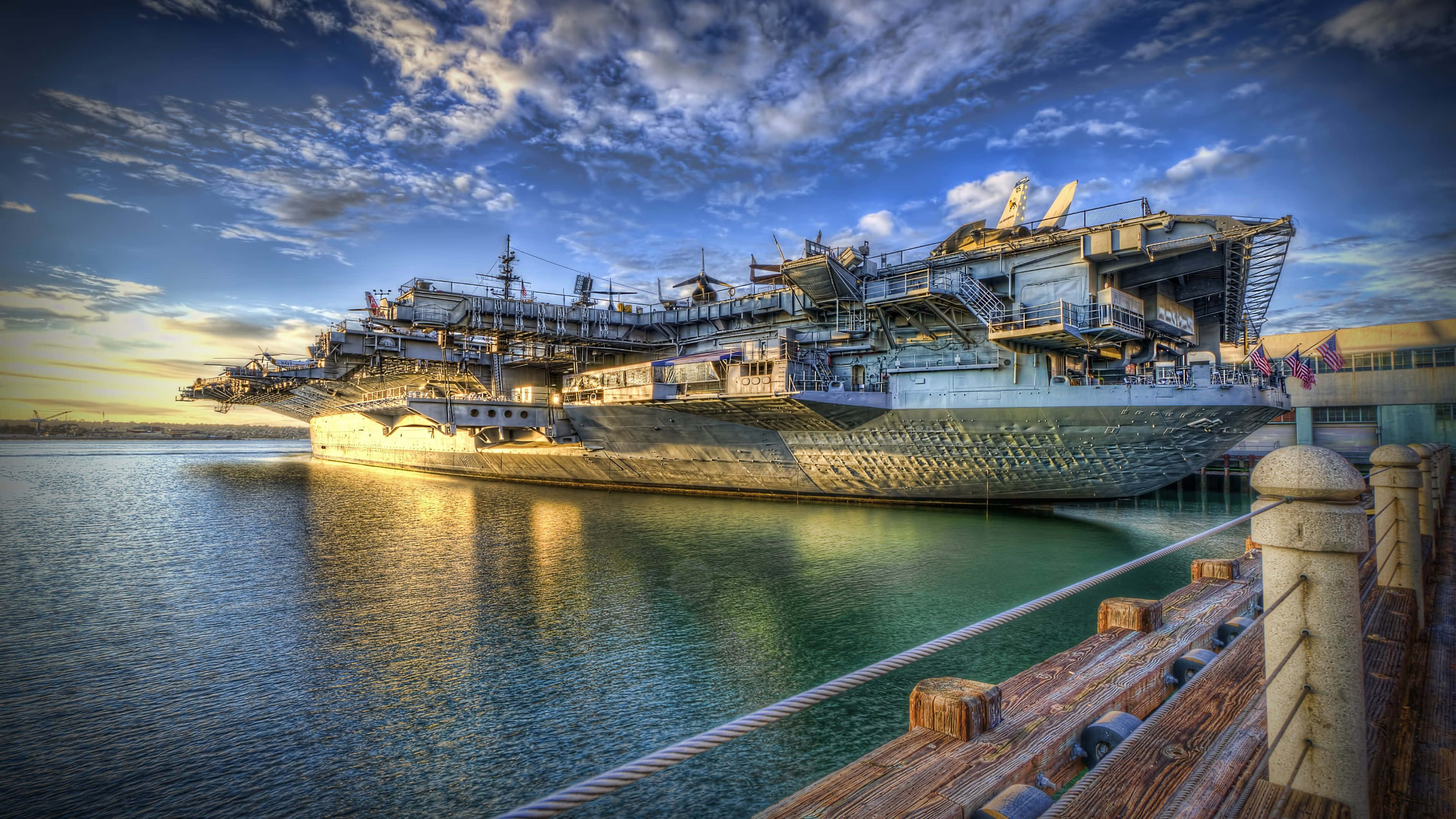 4K Hdr 桌布 壁紙 — 一艘巨大的航空母艦停泊在平靜的水域中，上方是帶有金色雲彩的壯麗日落天空。