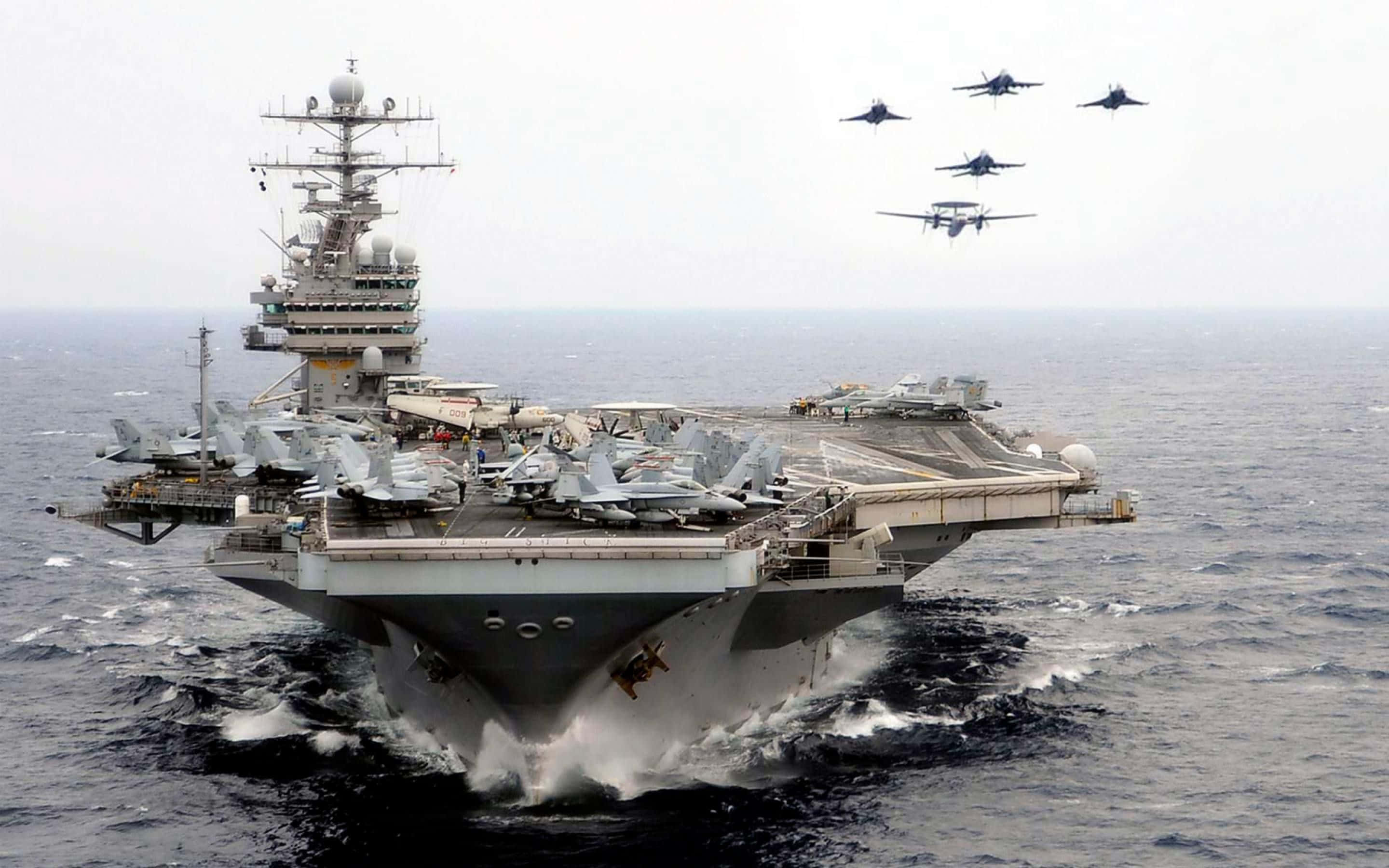 Navy 桌布 壁紙 — 一艘巨大的航空母艦在波濤洶湧的灰色海洋中航行，戰鬥機在上空編隊飛行。