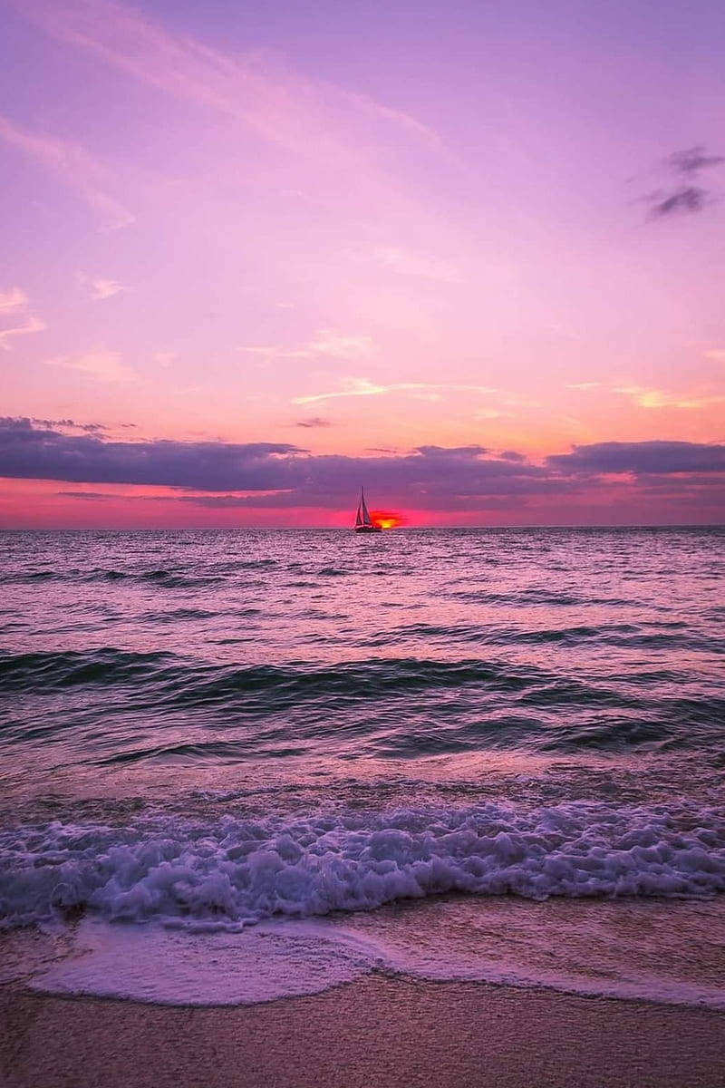 海灘 桌布 壁紙 — 一艘帆船在日落時分滑過寧靜的海洋，海浪在充滿活力的紫色和粉紅色天空下拍打著沙灘。