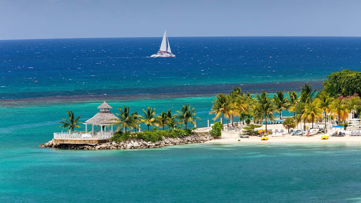 Jamaican Island 桌布 壁紙 — 一艘帆船在熱帶島嶼附近碧綠色的水面上滑行，島上有棕櫚樹和一個茅草涼亭。
