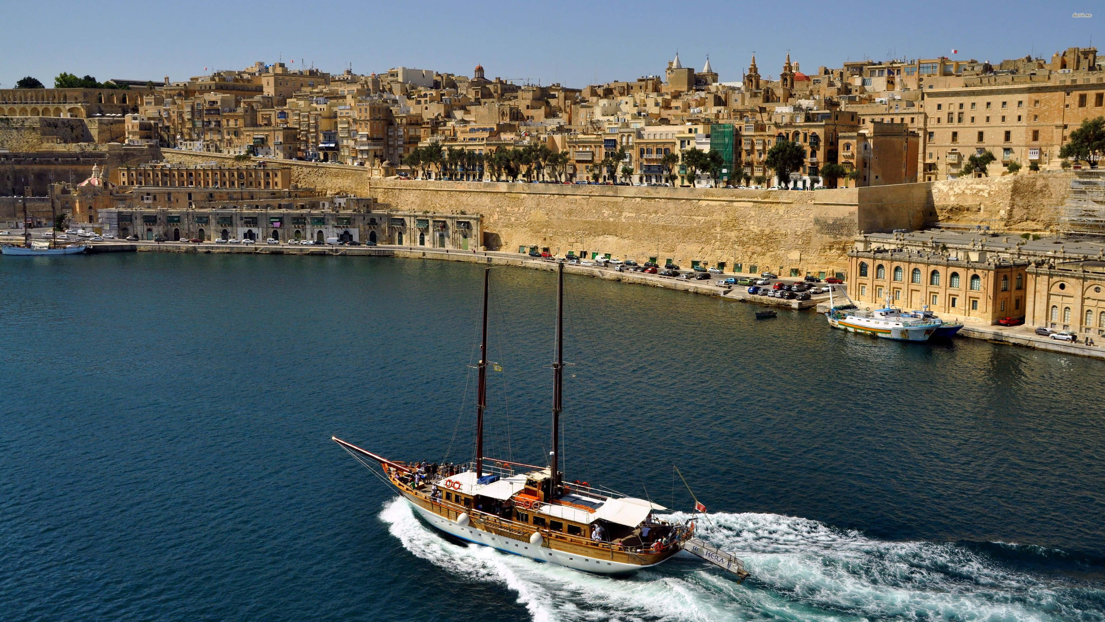 Malta 桌布 壁紙 — 一艘木製帆船在陽光普照的歷史海岸小鎮附近，劃過深藍色的海水，小鎮有陶土屋頂和石牆。