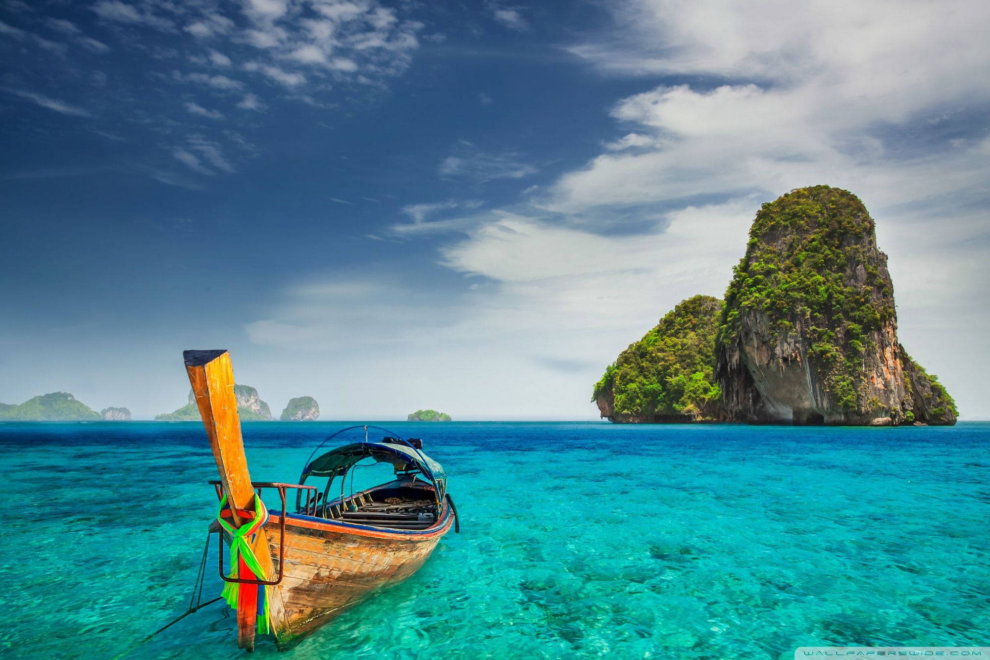 Boat 桌布 壁紙 — 一艘木製長尾船漂浮在清澈碧綠的海水中，靠近一個鬱鬱蔥蔥的綠色喀斯特島嶼，天空湛藍，雲朵稀疏。