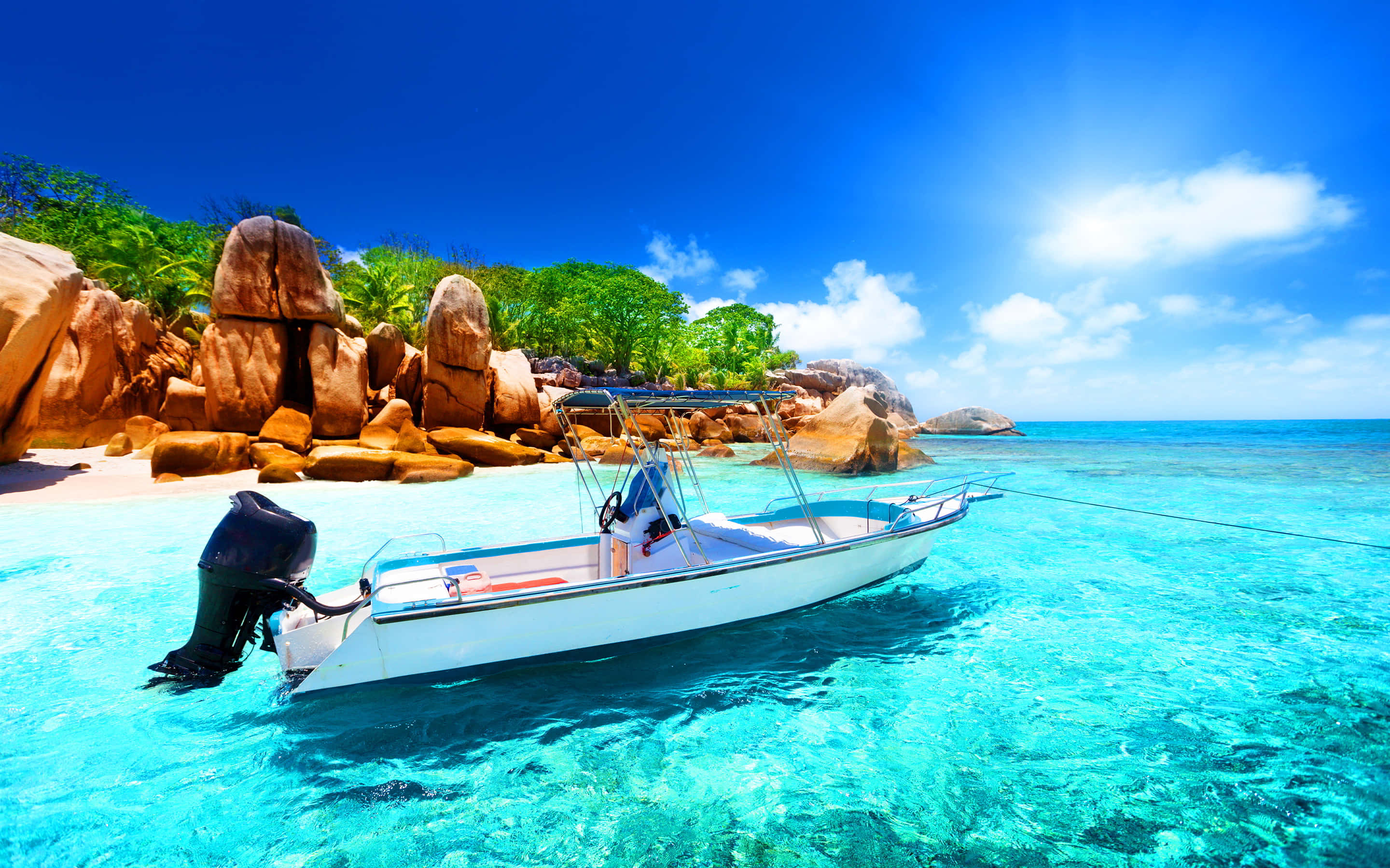 Seychelles Beach 桌布 壁紙 — 一艘白色機動船漂浮在清澈碧綠的海水中，旁邊是沙灘，沙灘被巨大的鏽色岩石和茂密的綠色植物環繞，上方是明亮的藍天。