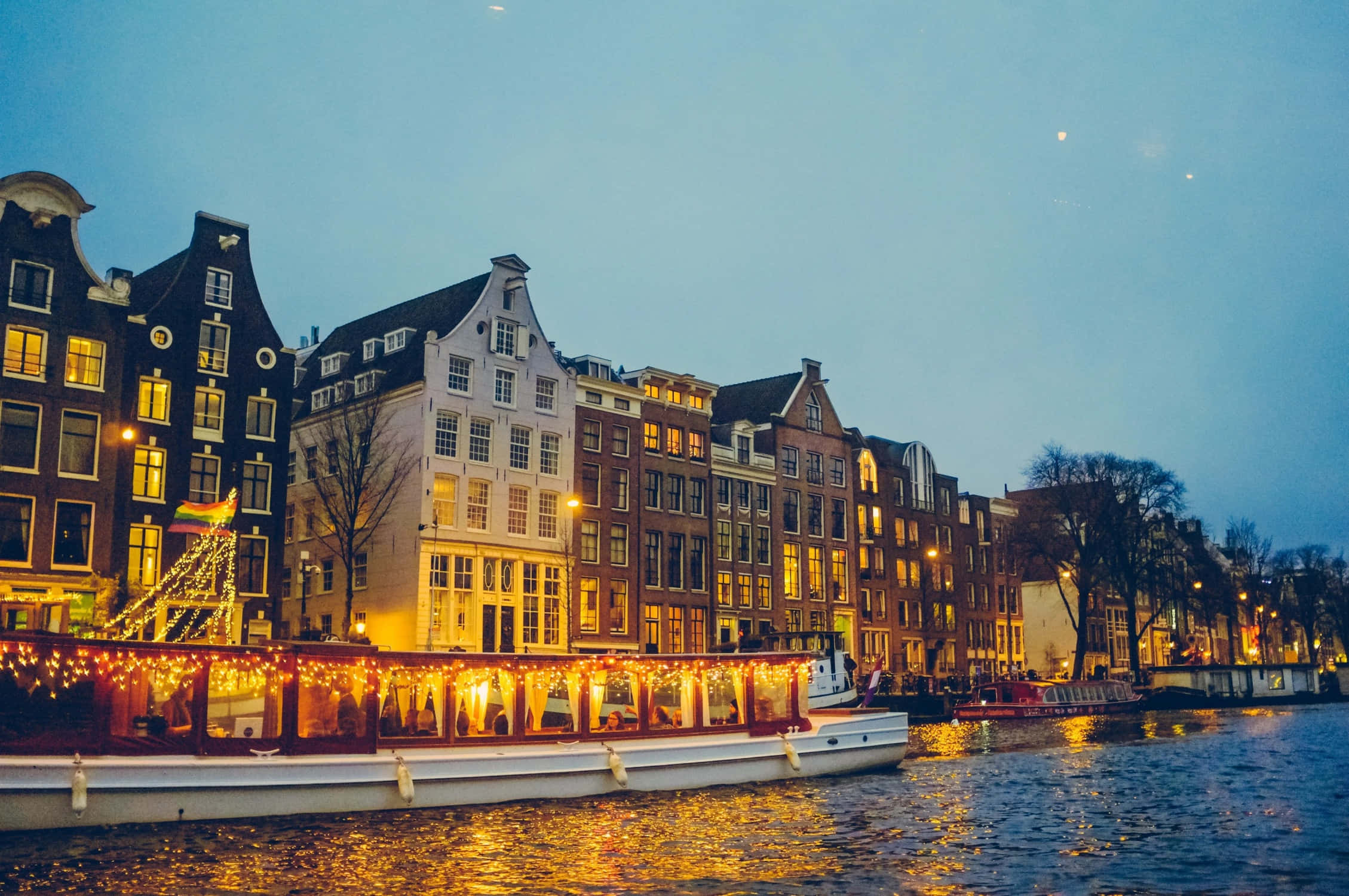 Netherlands 桌布 壁紙 — 一艘節日運河船裝飾著溫暖的串燈，在暮色中滑過閃爍的歷史荷蘭房屋。