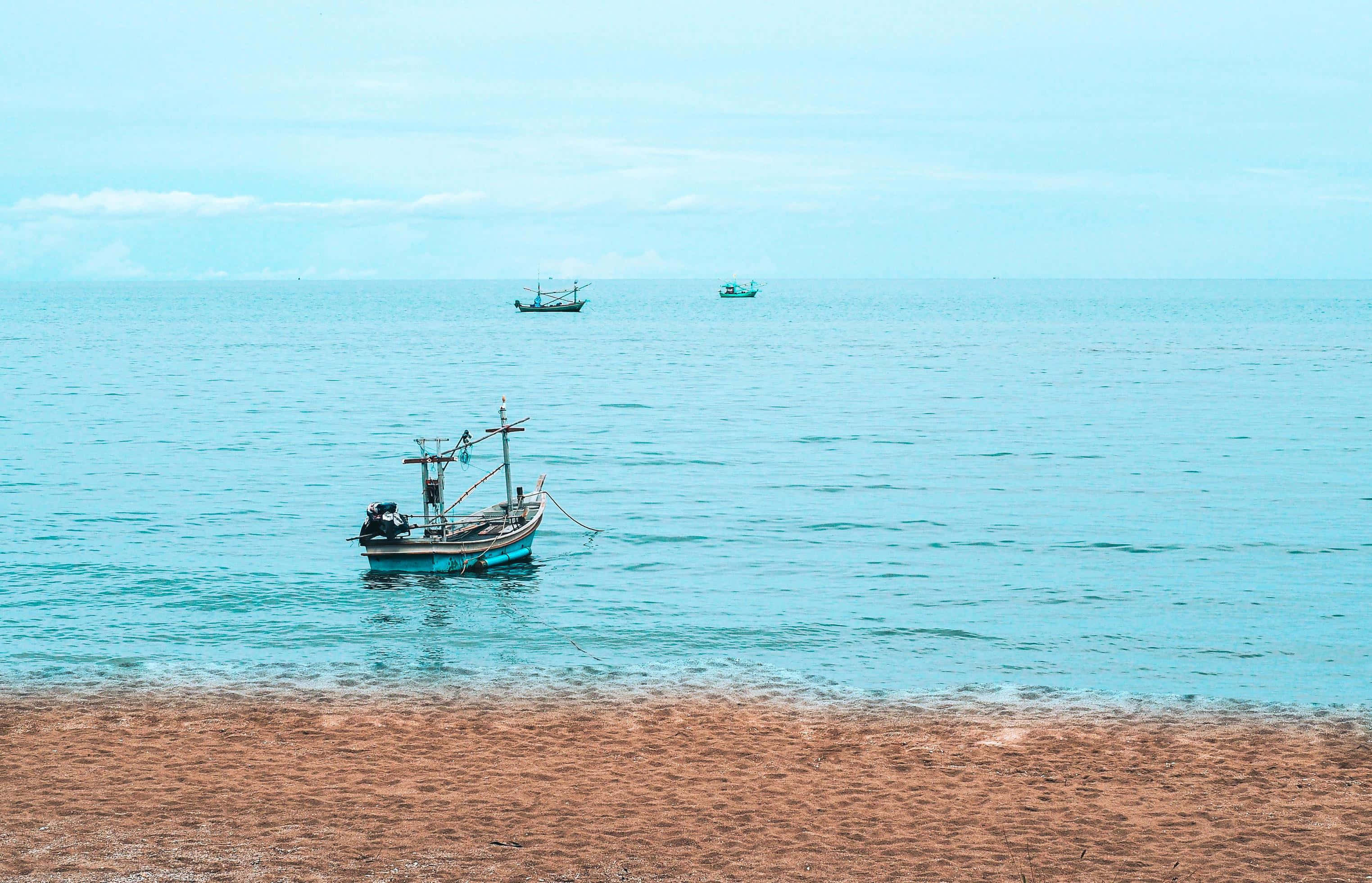 Boating 桌布 壁紙 — 一艘經風吹日曬的漁船停泊在沙灘附近，另外兩艘船在淡色天空下平靜地漂浮在碧綠色的水面上。