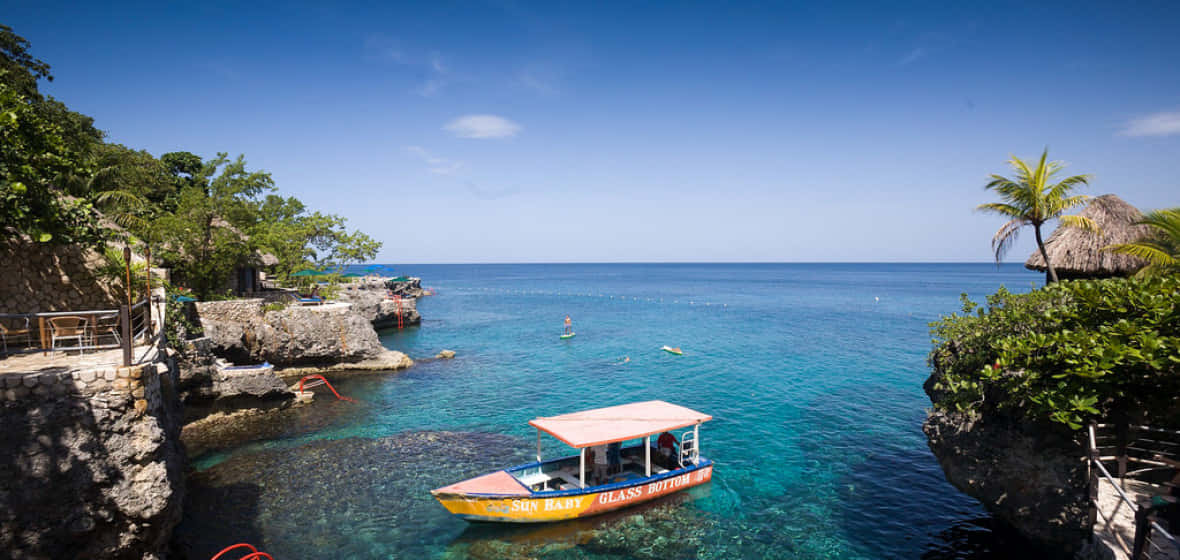 Jamaican Island 桌布 壁紙 — 一艘色彩鮮豔的玻璃底船在明亮的藍天下，穿梭於鬱蔥的綠色懸崖之間，滑行在清澈見底的碧綠海水上。