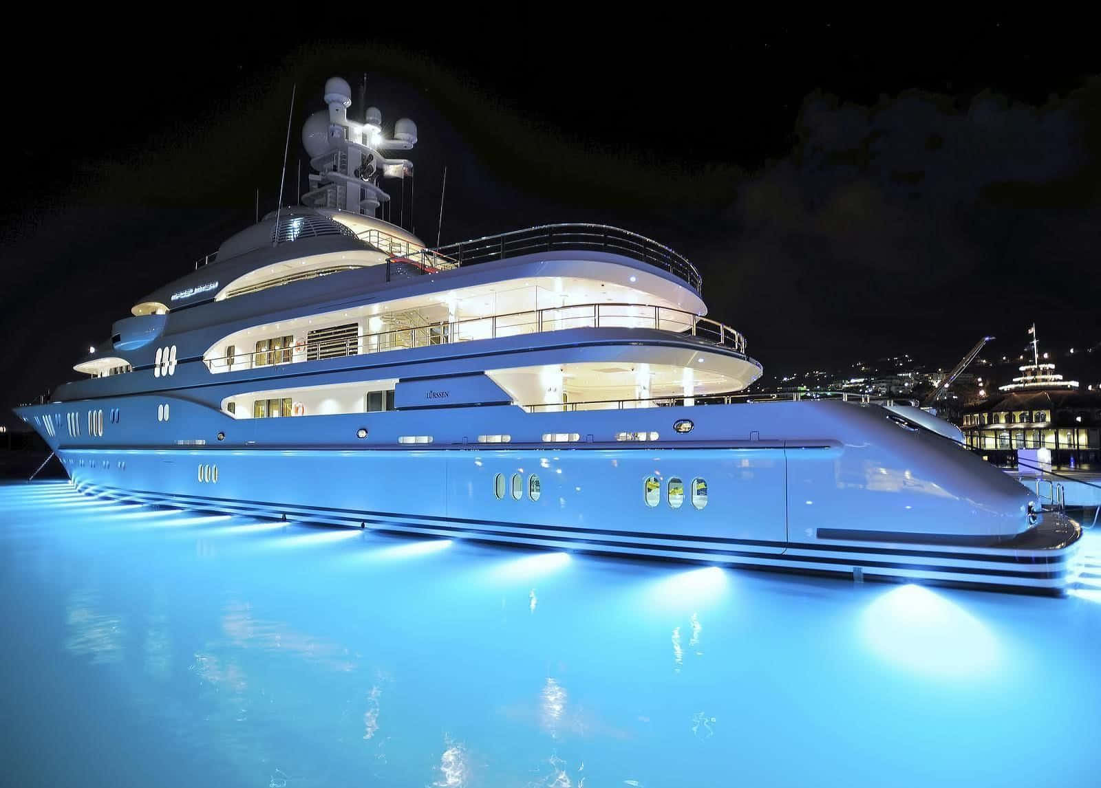 Yacht 桌布 壁紙 — 一艘豪華遊艇在夜間發出藍色光芒，其時尚的白色船體倒映在靠近城市天際線的平靜水面上。