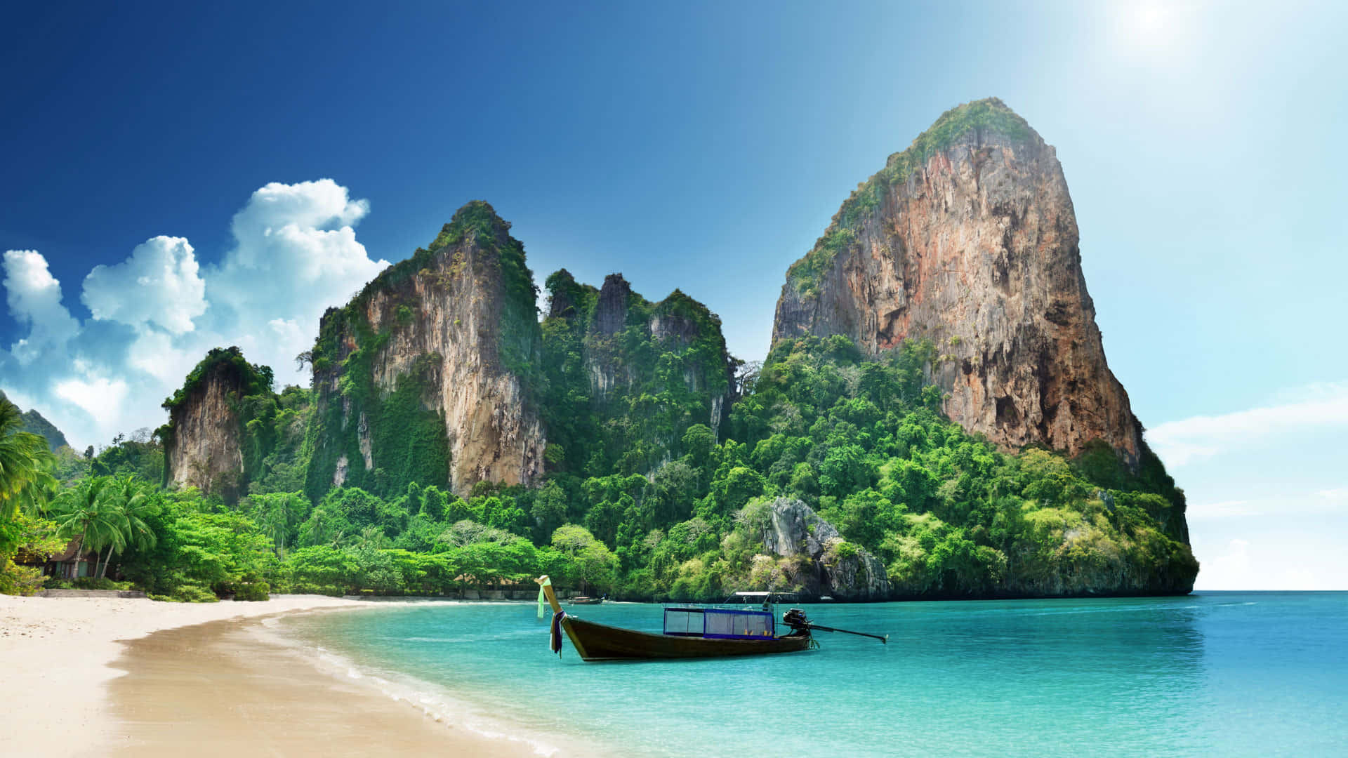 Beautiful Travel 桌布 壁紙 — 一艘長尾船停泊在碧綠色的海面上，旁邊是白色沙灘，四周被高聳的叢林覆蓋的石灰岩懸崖環繞，天空湛藍明亮。