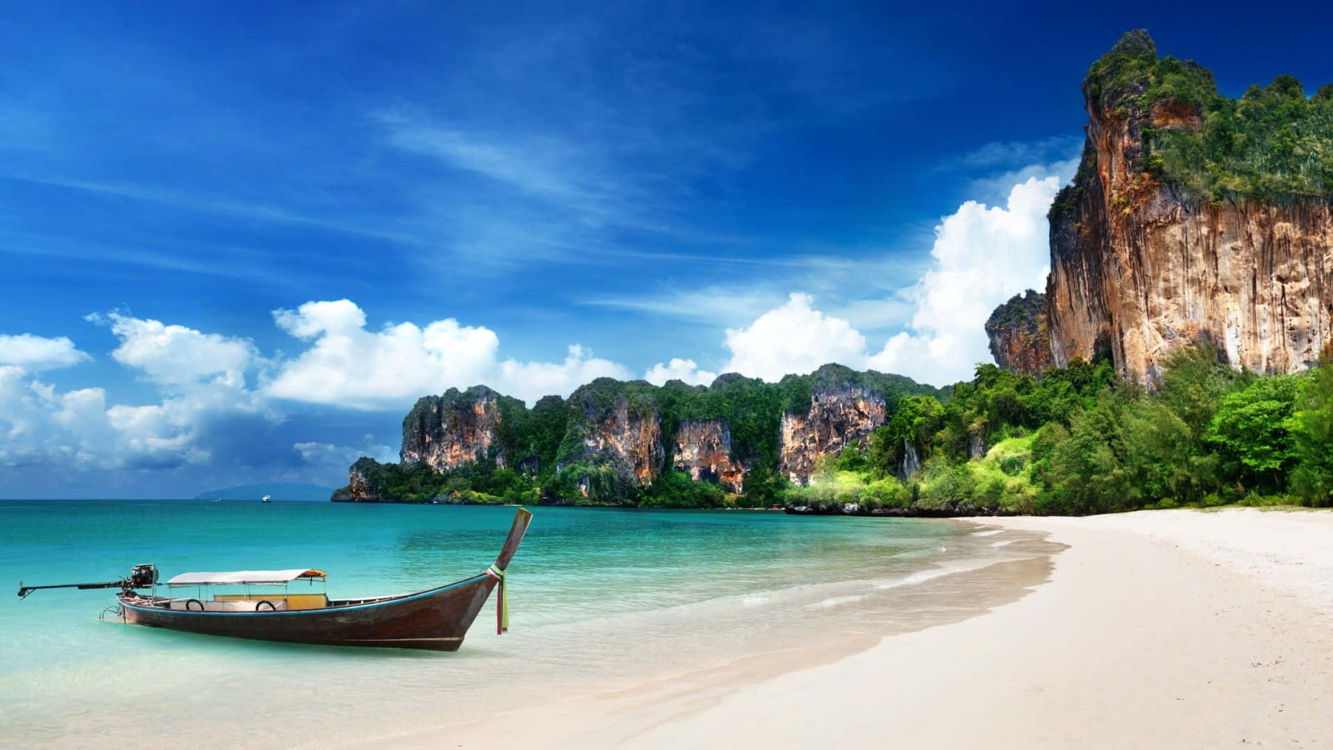 Thailand 桌布 壁紙 — 一艘長尾船停泊在純淨的白色沙灘上，旁邊是碧綠的海水，戲劇性的石灰岩懸崖在茂密的綠植後方升起，上方是明亮的藍天。