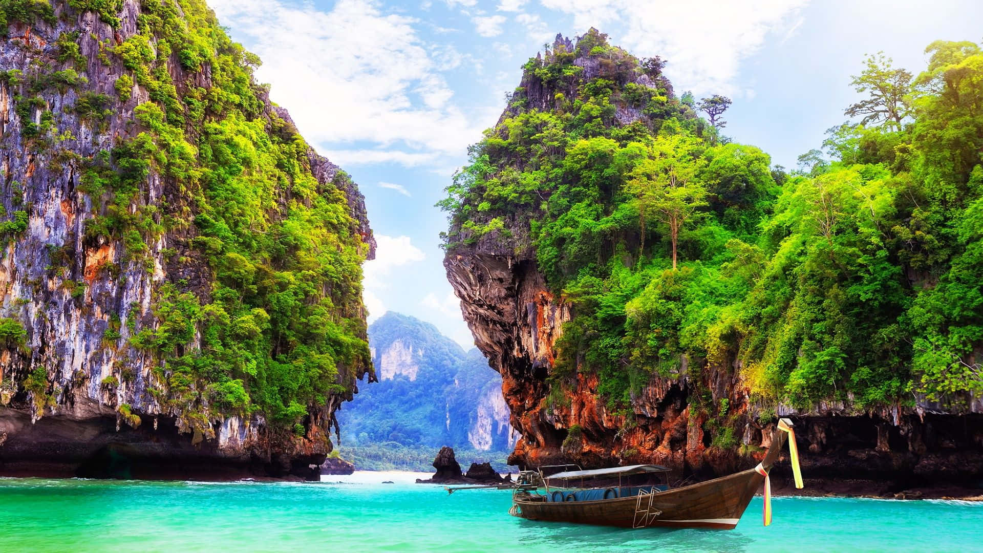 Thailand Beach 桌布 壁紙 — 一艘長尾船漂浮在碧綠色的海水上，兩旁是聳立的叢林覆蓋的石灰岩懸崖，上方是明亮的藍天。