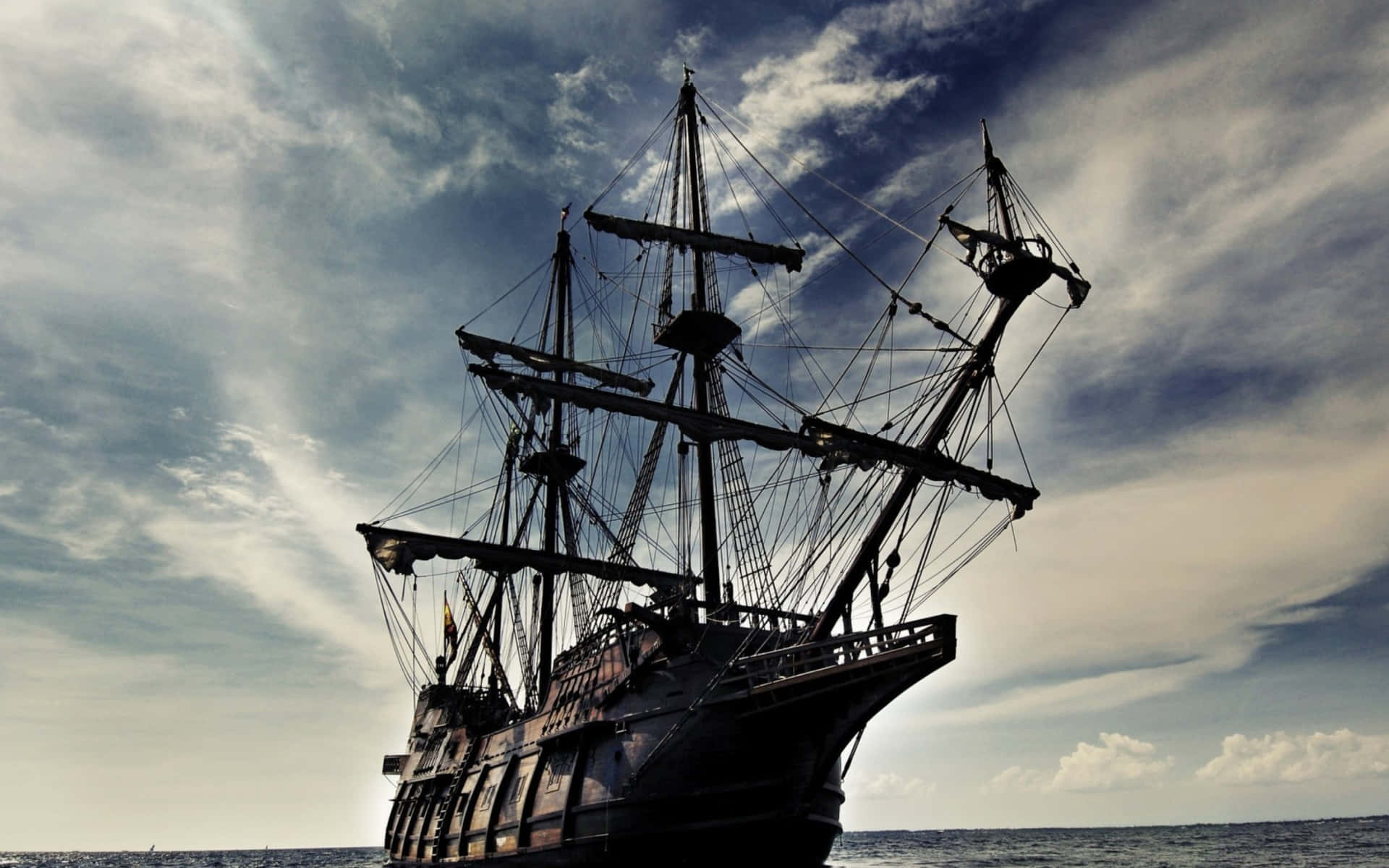 Pirates Of The Caribbean 桌布 壁紙 — 一艘雄偉的海盜船在戲劇性的多雲天空下航行，帆布收攏，駛向開闊的海洋。