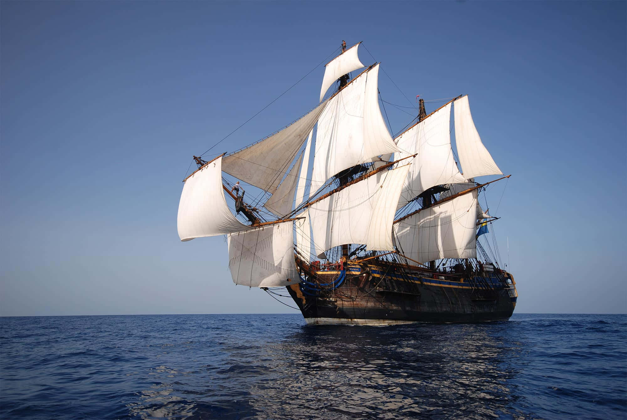 Ship 桌布 壁紙 — 一艘雄偉的高桅帆船，鼓滿白色風帆，在晴朗的天空下滑過平靜的藍色水域。