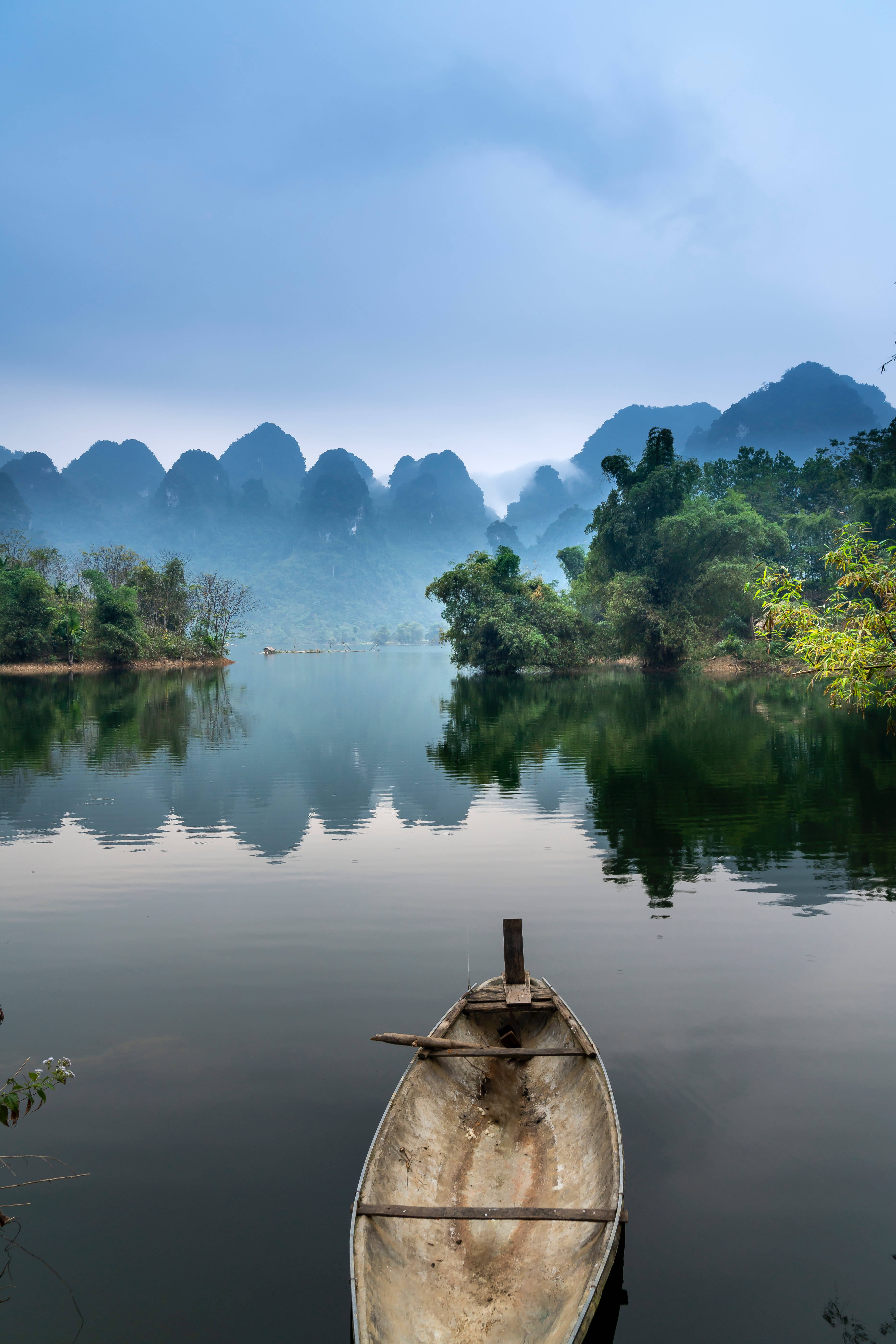 river 桌布 壁紙 — 一艘風化的木船在霧氣瀰漫的河流上靜靜漂浮，映照著喀斯特山脈，天空柔和湛藍。