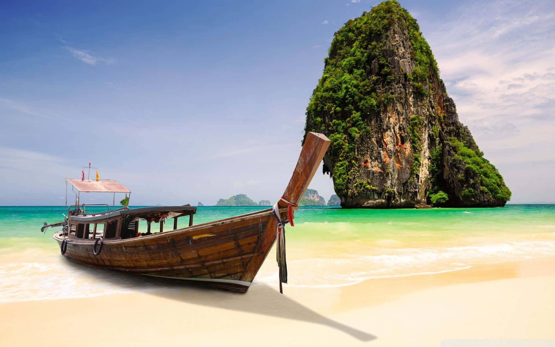 Thailand 桌布 壁紙 — 一艘風化的木製長尾船停泊在純淨的白色沙灘上，旁邊是碧綠的海水，背景是一座高聳的、被叢林覆蓋的石灰岩島嶼。