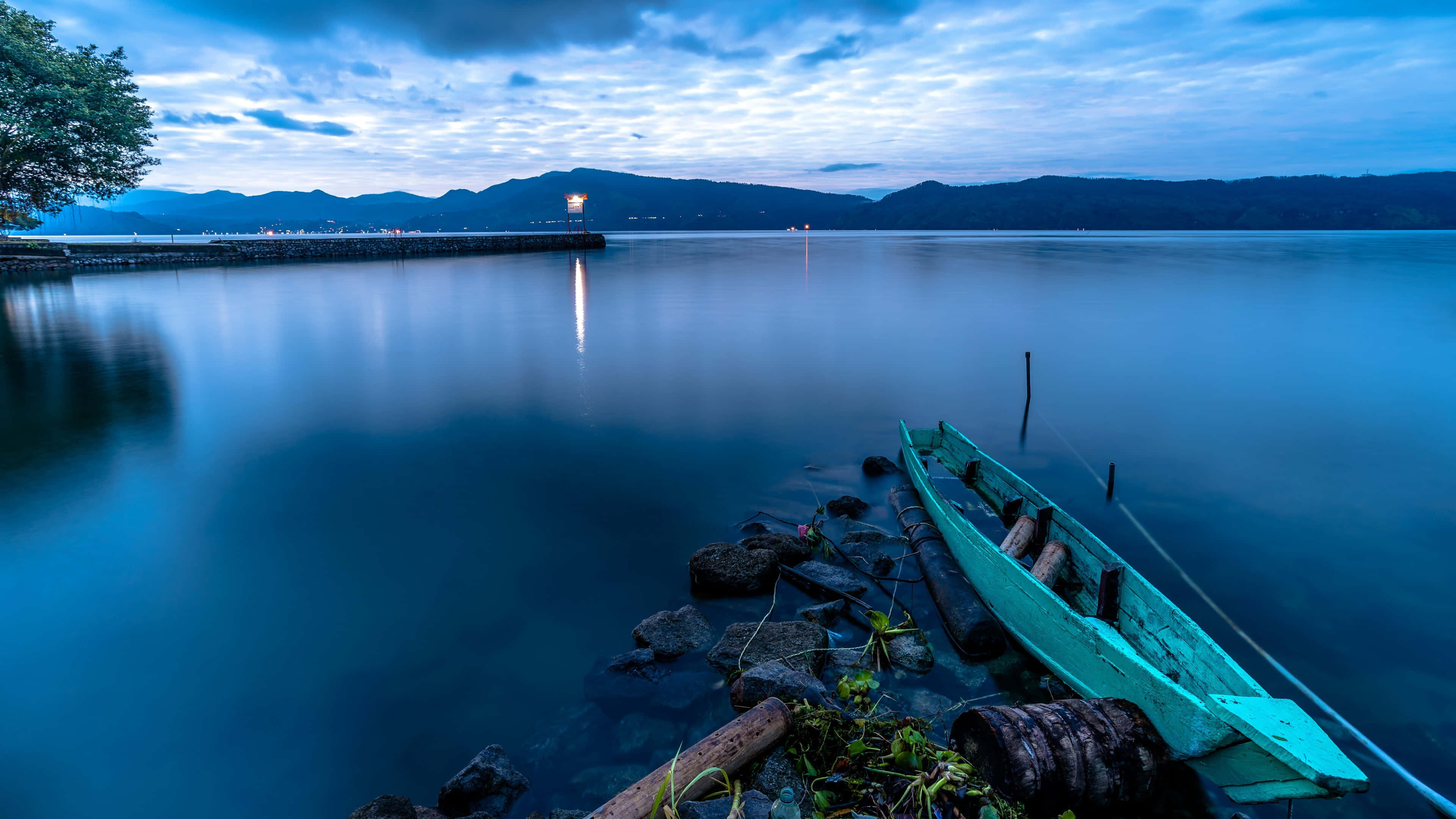 Indonesia 桌布 壁紙 — 一艘風化的藍綠色小船在黃昏時停靠在岩石海岸上，平靜的藍色水面倒映著遠處的燈光和山脈。