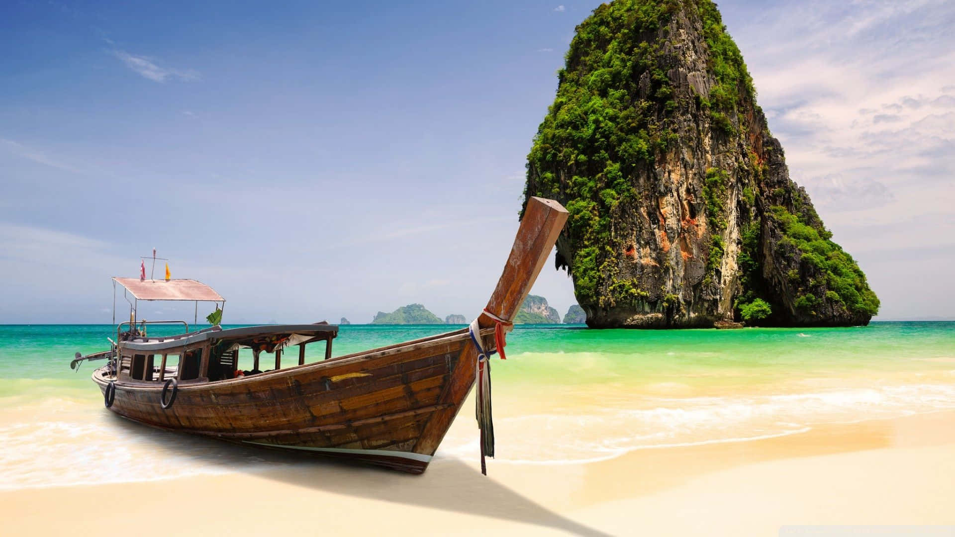 Thailand Beach 桌布 壁紙 — 一艘飽經風霜的木製長尾船停泊在原始白色沙灘上，旁邊是碧綠的海水，背景中一座鬱鬱蔥蔥的綠色喀斯特島嶼戲劇性地聳立。