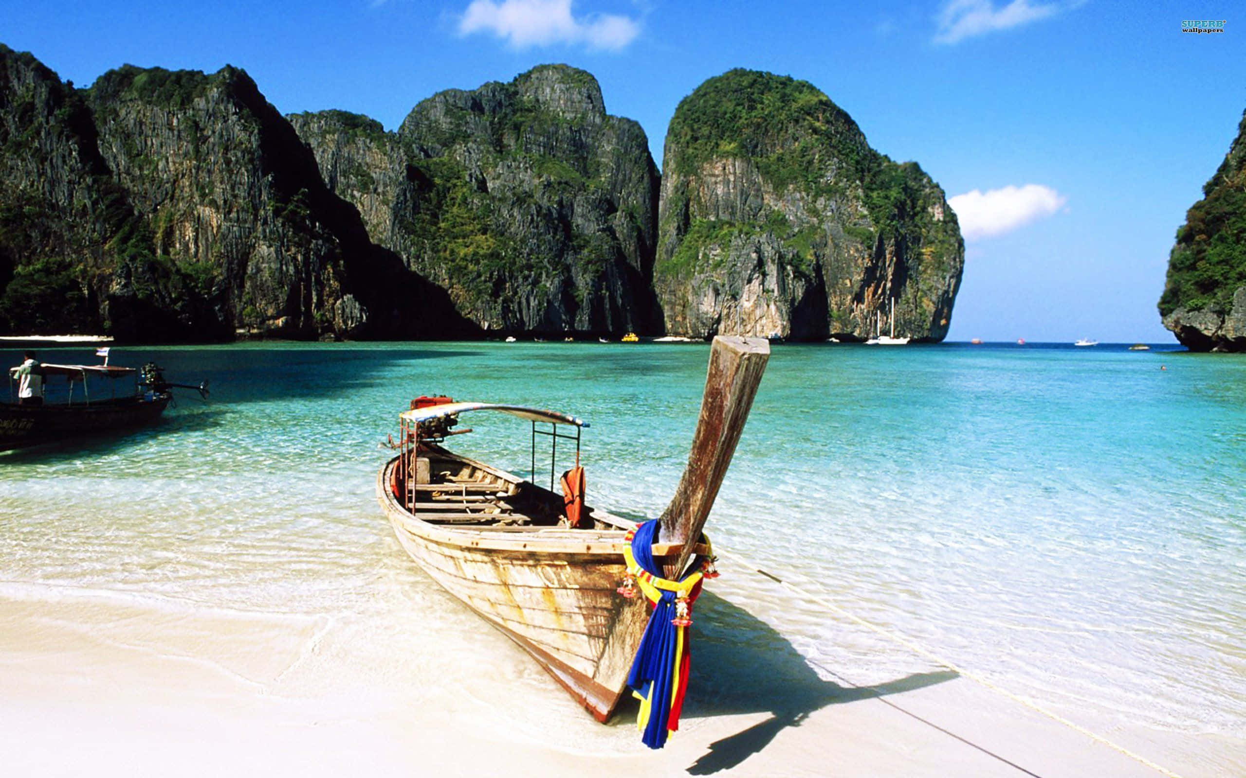 Thailand 桌布 壁紙 — 一艘飽經風霜的木製長尾船停泊在純淨的白色沙灘上，周圍是碧綠的海水、高聳的叢林覆蓋的石灰岩懸崖，頭頂是明亮的藍天。