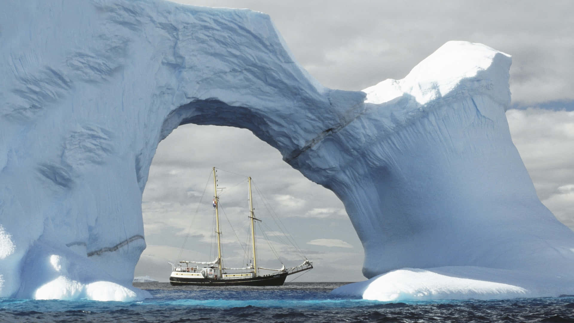 Antarctica 桌布 壁紙 — 一艘高桅帆船在陰天之下，穿越南極冰冷海域中一個巨大的藍色冰拱門。