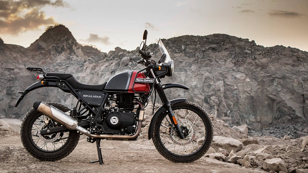 自行車 桌布 壁紙 — 一輛 Royal Enfield Himalayan 電單車在崎嶇地形上蓄勢待發，背景是岩石山脈，天空呈現柔和的金色。