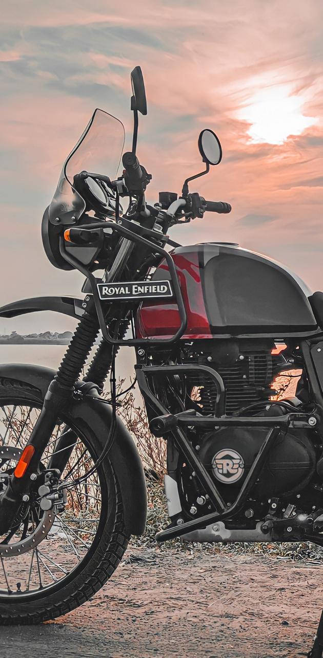 自行車 桌布 壁紙 — 一輛 Royal Enfield 電單車在日落時分停在碎石上，背後是溫暖的粉紅色和橙色天空。