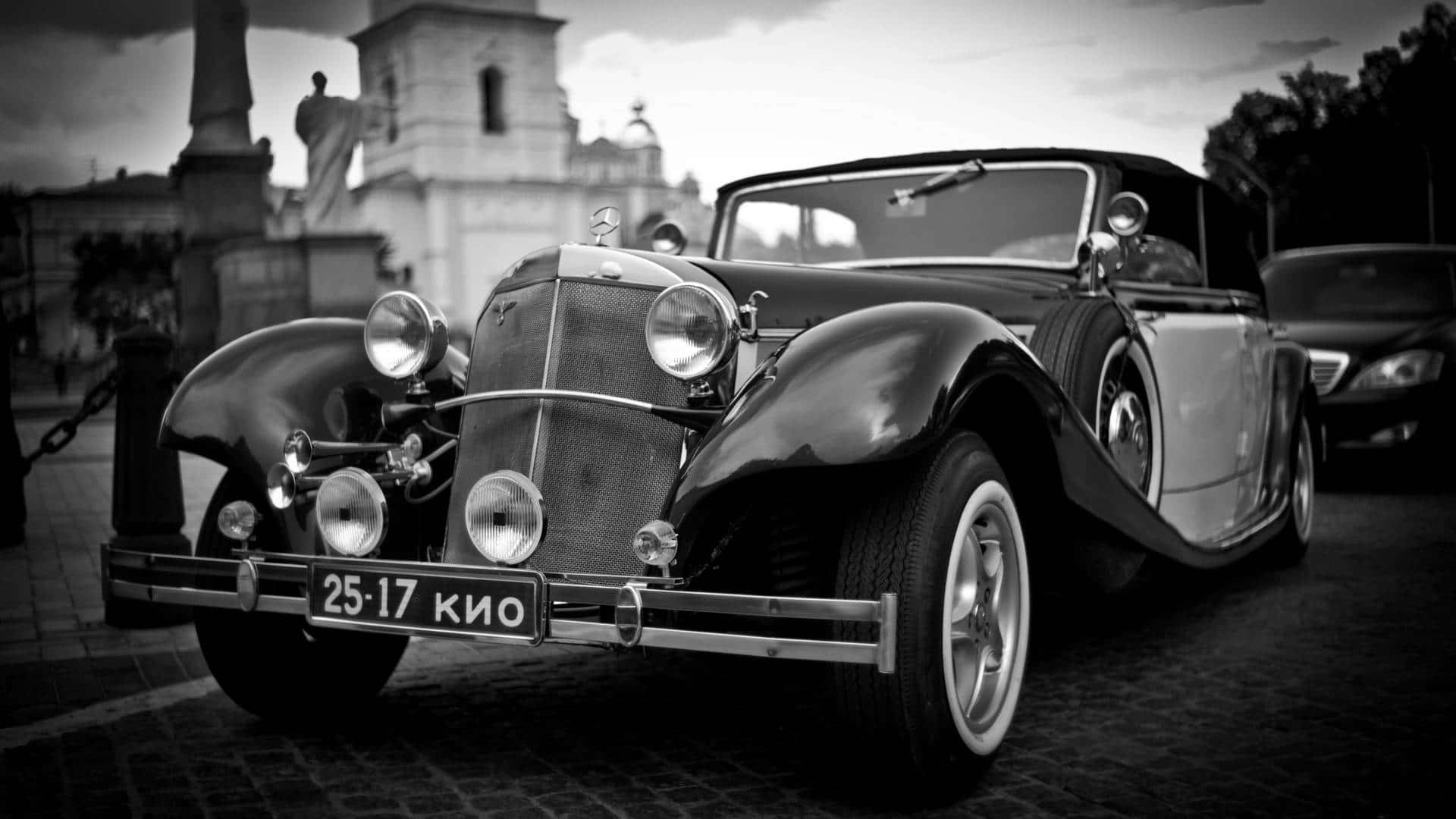 Black And White Vintage Car 桌布 壁紙 — 一輛 vintage Mercedes-Benz 開篷車停泊在鵝卵石街道上，背景是歷史建築。