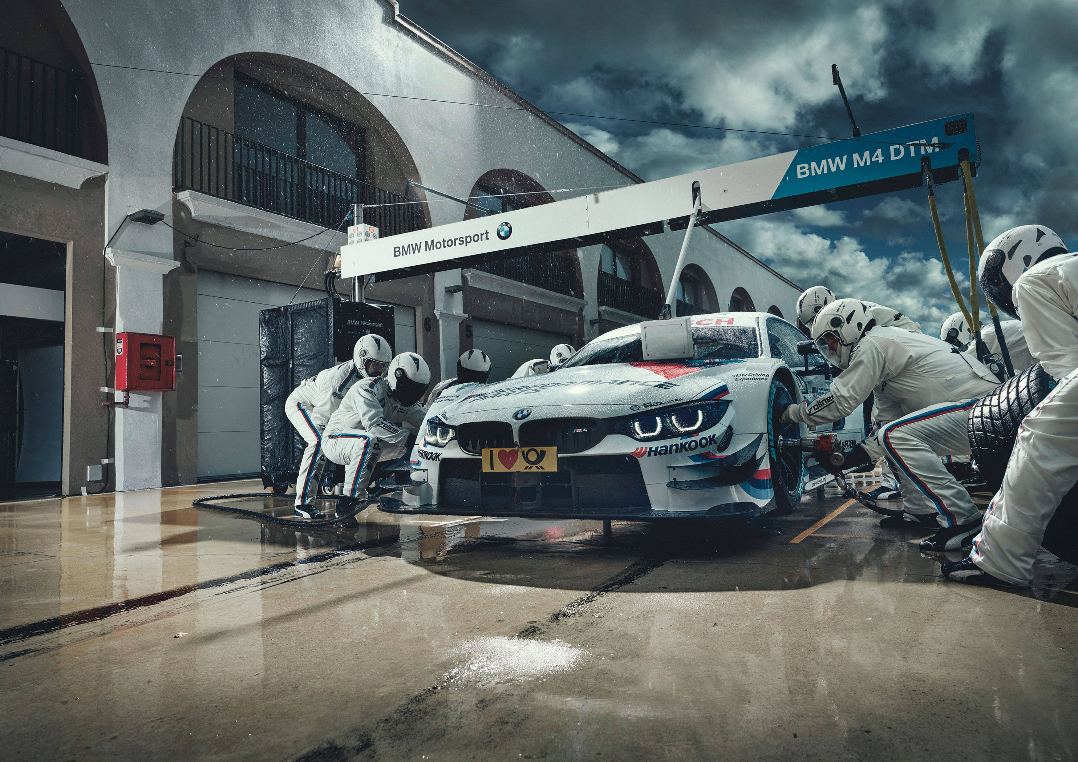 Sports Car 桌布 壁紙 — 一輛BMW M4 DTM賽車正在暴風雨的天空下，在潮濕的車庫中由穿著白色制服的工作人員進行維修。