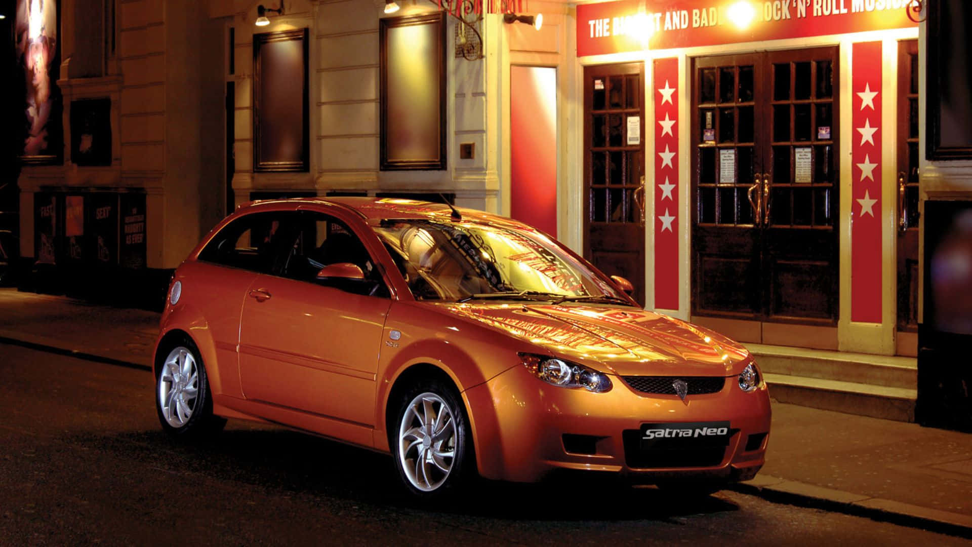Proton 桌布 壁紙 — 一輛亮橙色 Saturn Neo 掀背車在夜間停泊在一個燈火通明、帶有紅色星柱的搖滾博物館前。