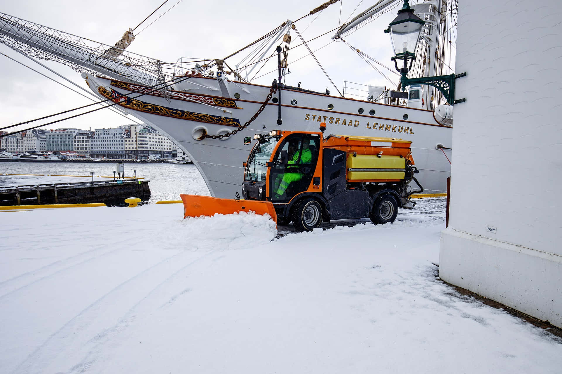 Snowplow 桌布 壁紙 — 一輛亮橙色的掃雪機在雪港停泊的歷史白色船隻Statsraad Lehmkuhl旁邊清除積雪。