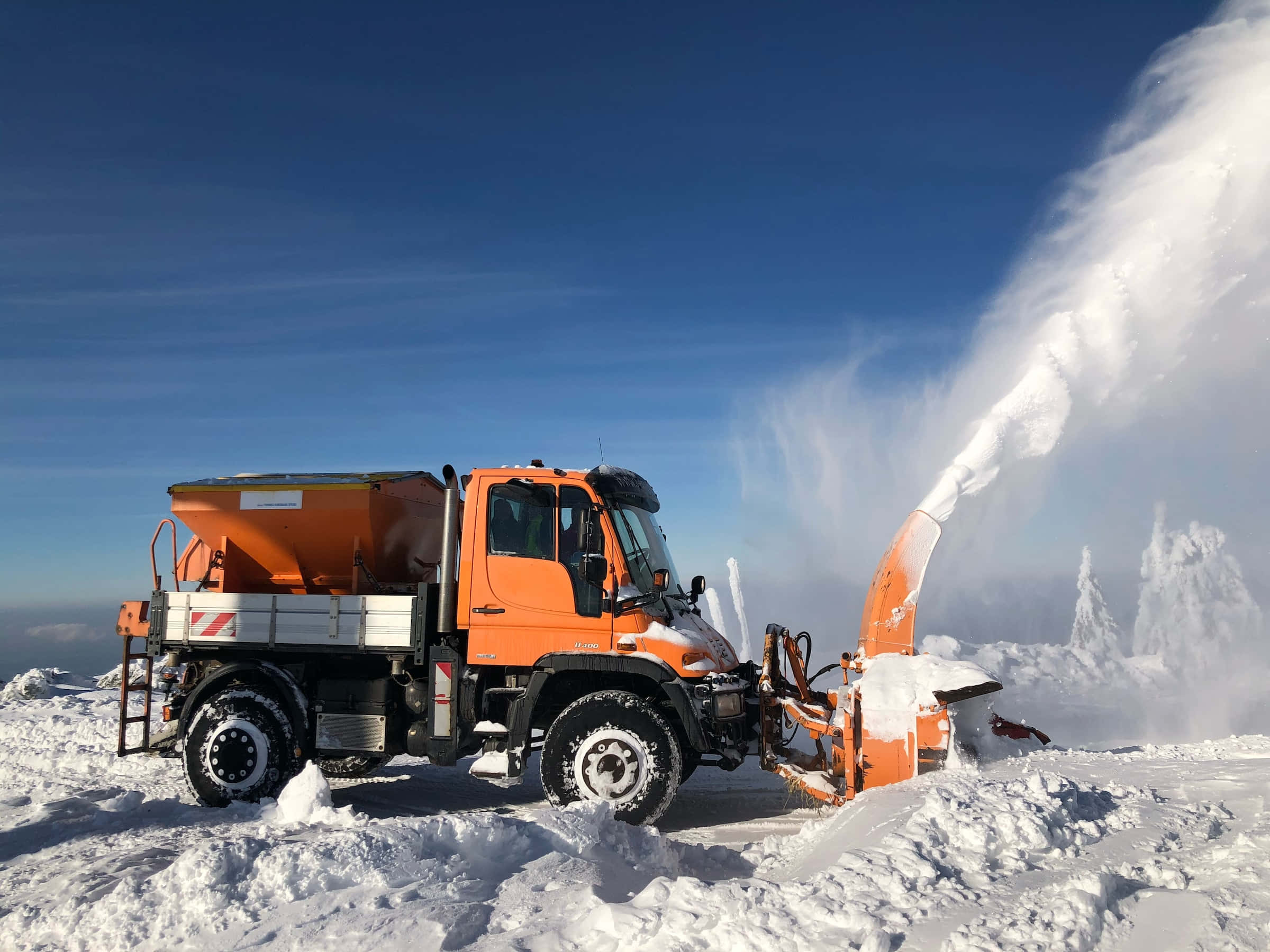 Snowplow 桌布 壁紙 — 一輛亮橙色的除雪卡車在白雪皚皚的山頂上，對著清澈的藍天將雪吹成旋轉的雲霧。