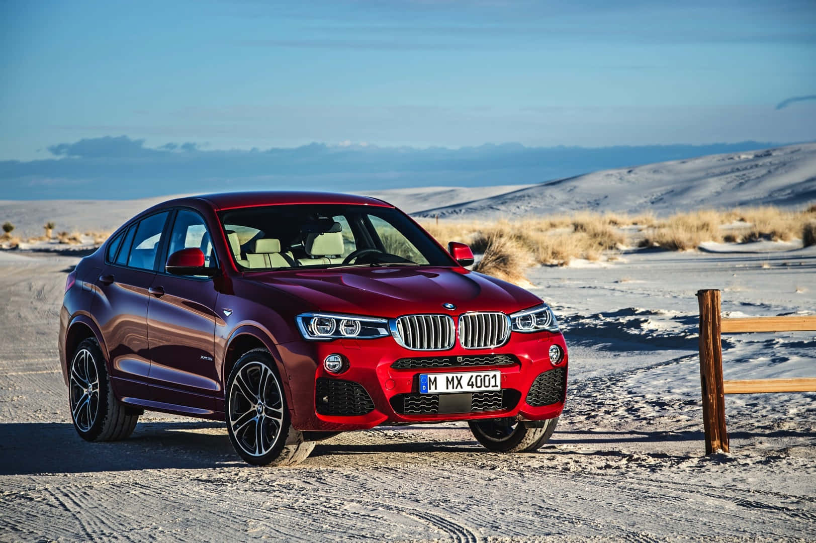 Bmw X4 桌布 壁紙 — 一輛亮紅色BMW X4 M停泊在白雪覆蓋的沙漠道路上，天空清澈湛藍，遠處有沙丘。