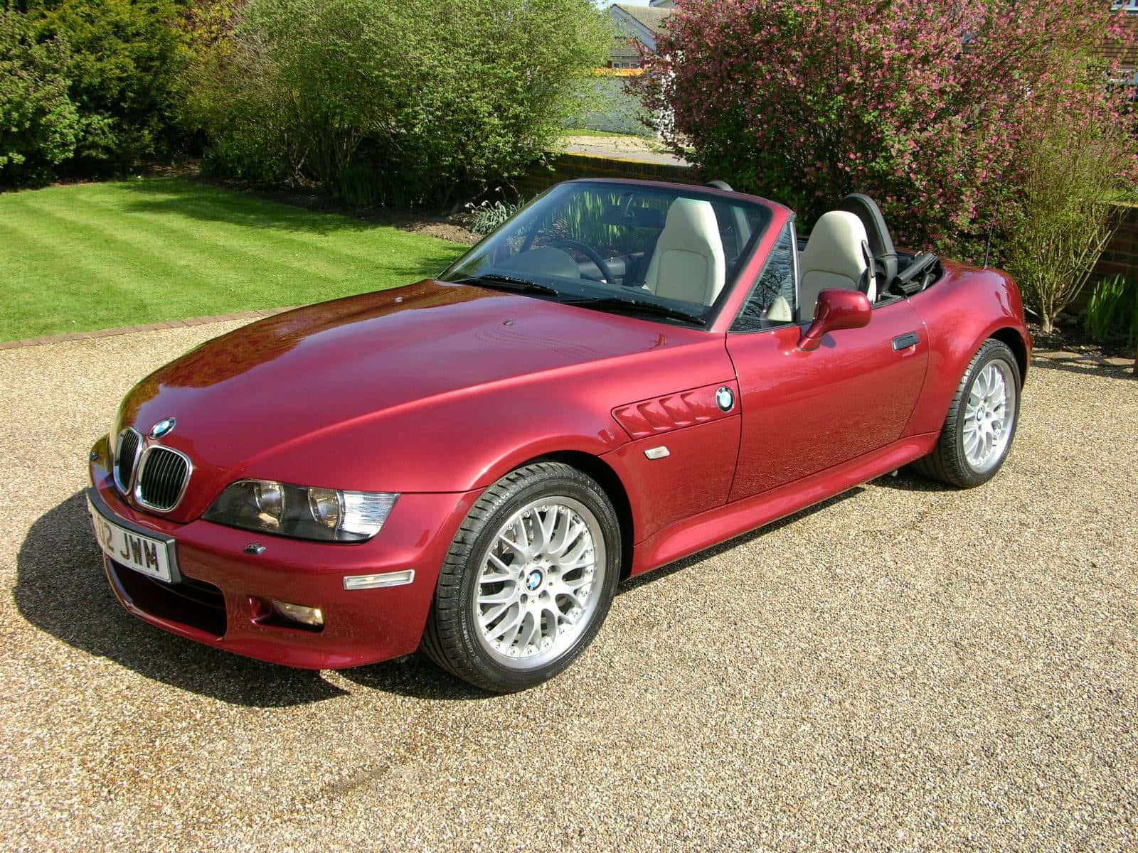 Bmw Z3 桌布 壁紙 — 一輛亮紅色BMW Z3開篷跑車停在碎石路上，周圍有綠色草坪和開花的灌木叢，在明亮的日光下。