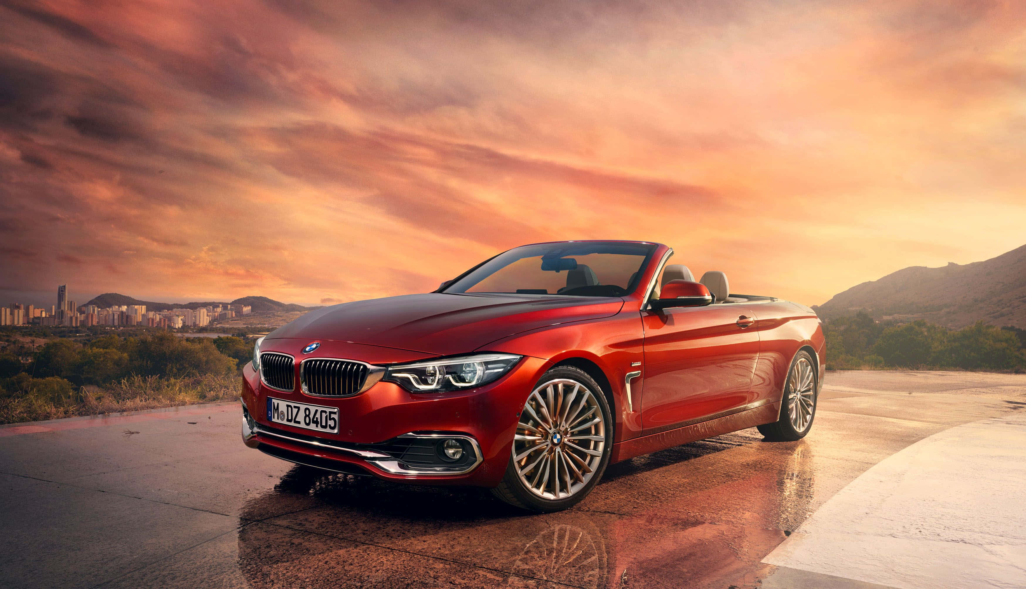 Bmw 4 Series 桌布 壁紙 — 一輛亮紅色BMW開篷車停泊在日落時分的濕滑道路上，背景是城市景觀和山脈。