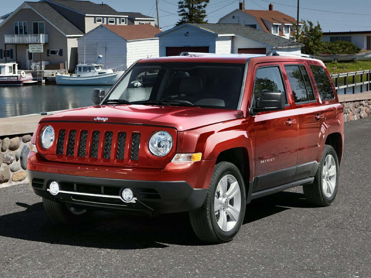 Jeep Patriot 桌布 壁紙 — 一輛亮紅色Jeep Patriot SUV停泊在瀝青路上，背景是陽光普照的海濱，有船隻和房屋。