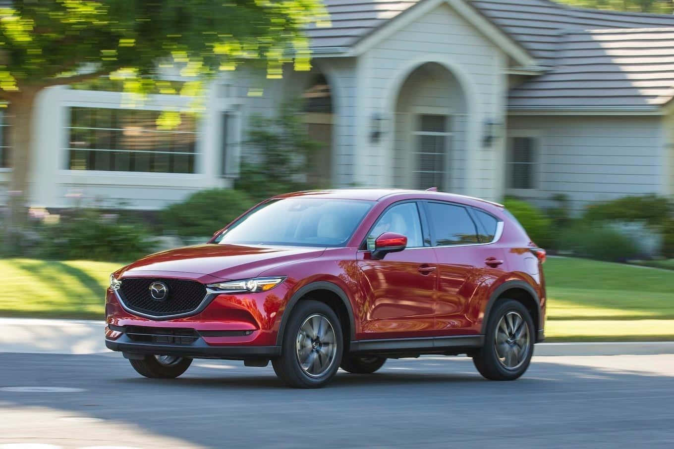 Mazda Cx-5 桌布 壁紙 — 一輛亮紅色Mazda CX-5 SUV在明亮的日光下駛過一棟帶有綠色草坪的白色郊區房屋。