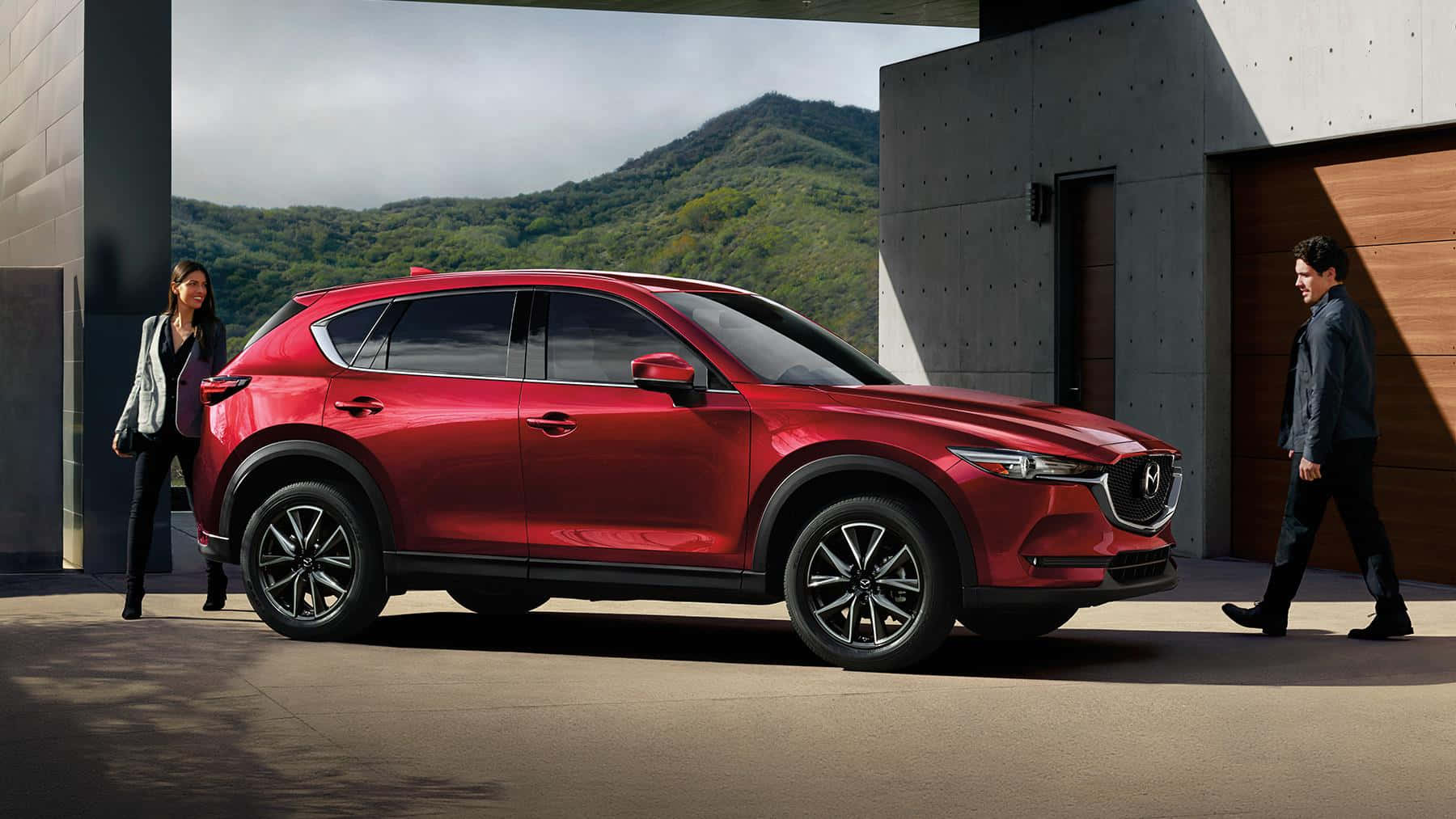 Mazda Cx-5 桌布 壁紙 — 一輛亮紅色SUV停在一棟現代混凝土建築前，附近有兩個人站立，背景是綠色山丘。