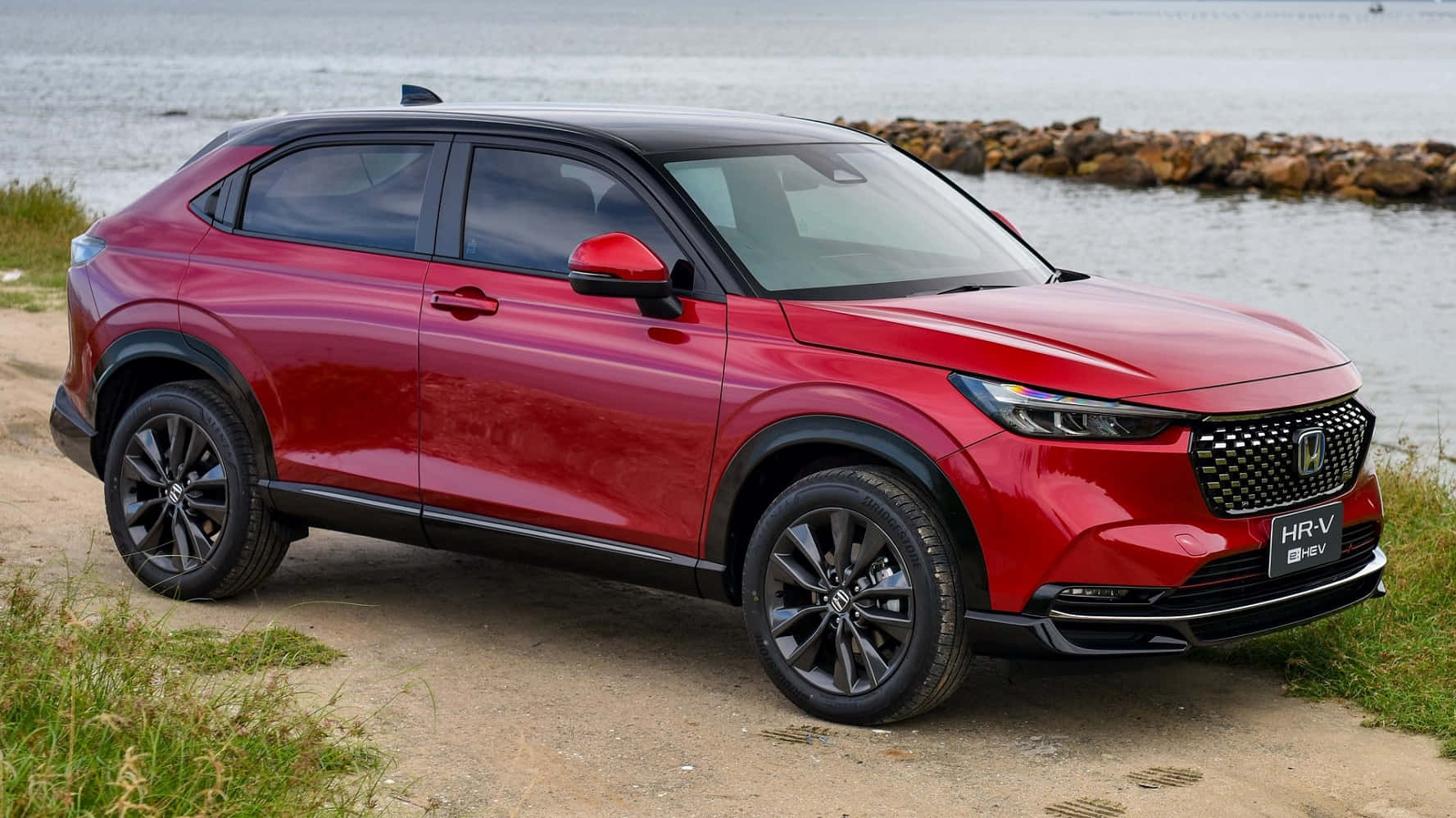 Honda Hr-v 桌布 壁紙 — 一輛亮紅色本田 HR-V 混合動力 SUV 停泊在草地岸邊，旁邊是平靜的水面，背景有岩石防波堤。