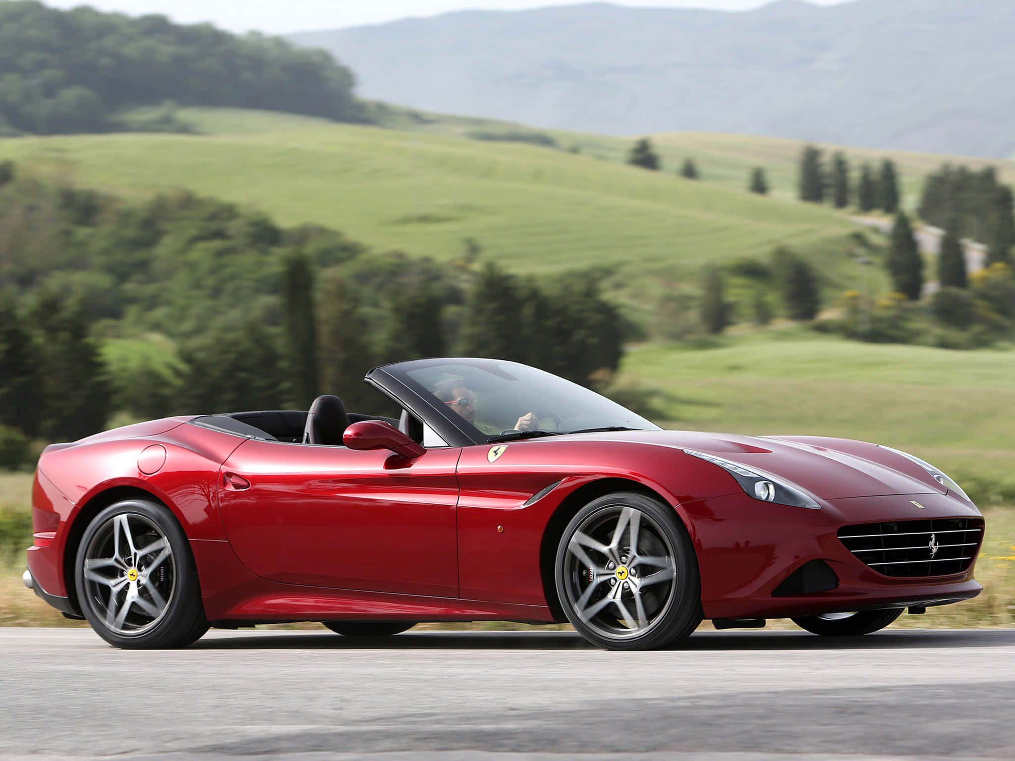 Ferrari California T 桌布 壁紙 — 一輛亮紅色法拉利California開篷車在明亮的日光下沿著蜿蜒的道路穿過鬱鬱蔥蔥的綠色山丘行駛。