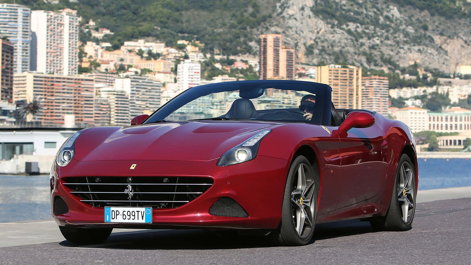 Ferrari California T 桌布 壁紙 — 一輛亮紅色法拉利California敞篷車停泊在沿海公路上，背景是摩納哥的天際線和山脈。