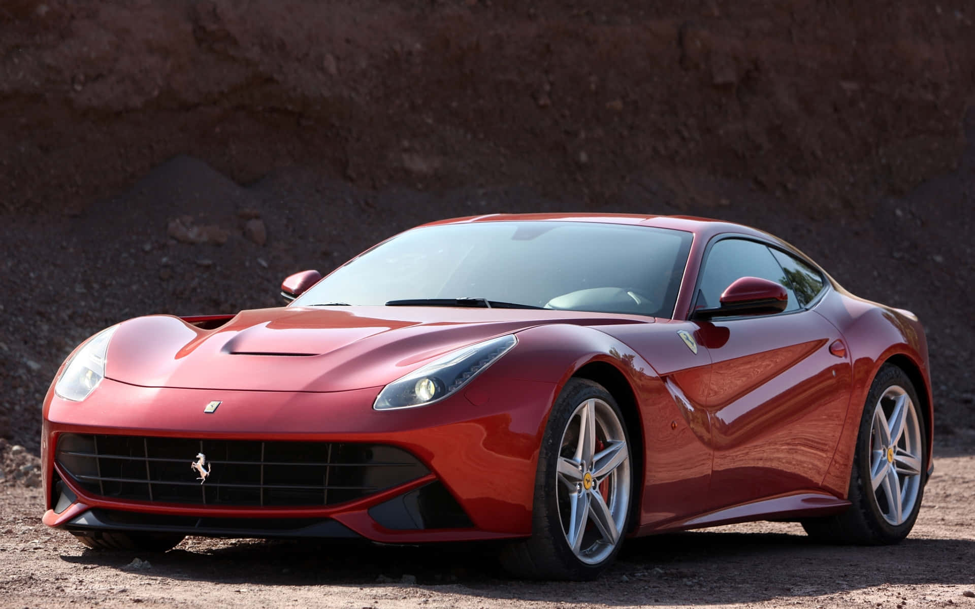 Ferrari F12 Berlinetta 桌布 壁紙 — 一輛亮紅色法拉利跑車停在碎石上，背景是崎嶇的岩石懸崖，在明亮的日光下。