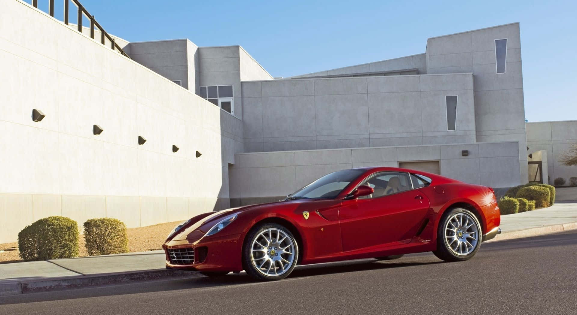 Ferrari 599 Gtb Fiorano 桌布 壁紙 — 一輛亮紅色法拉利跑車停泊在街道上，旁邊是現代混凝土建築，天空清澈湛藍。