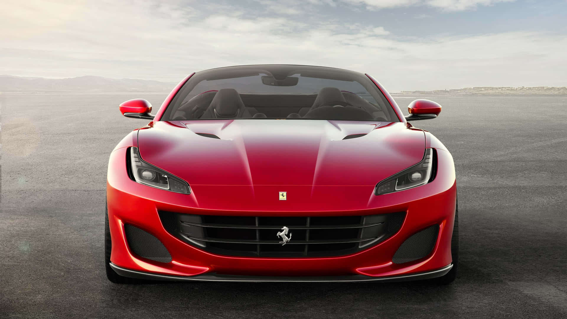 Ferrari Portofino 桌布 壁紙 — 一輛亮紅色法拉利跑車在柔和的陰天天空下，面向前方停放在廣闊的灰色瀝青路面上。