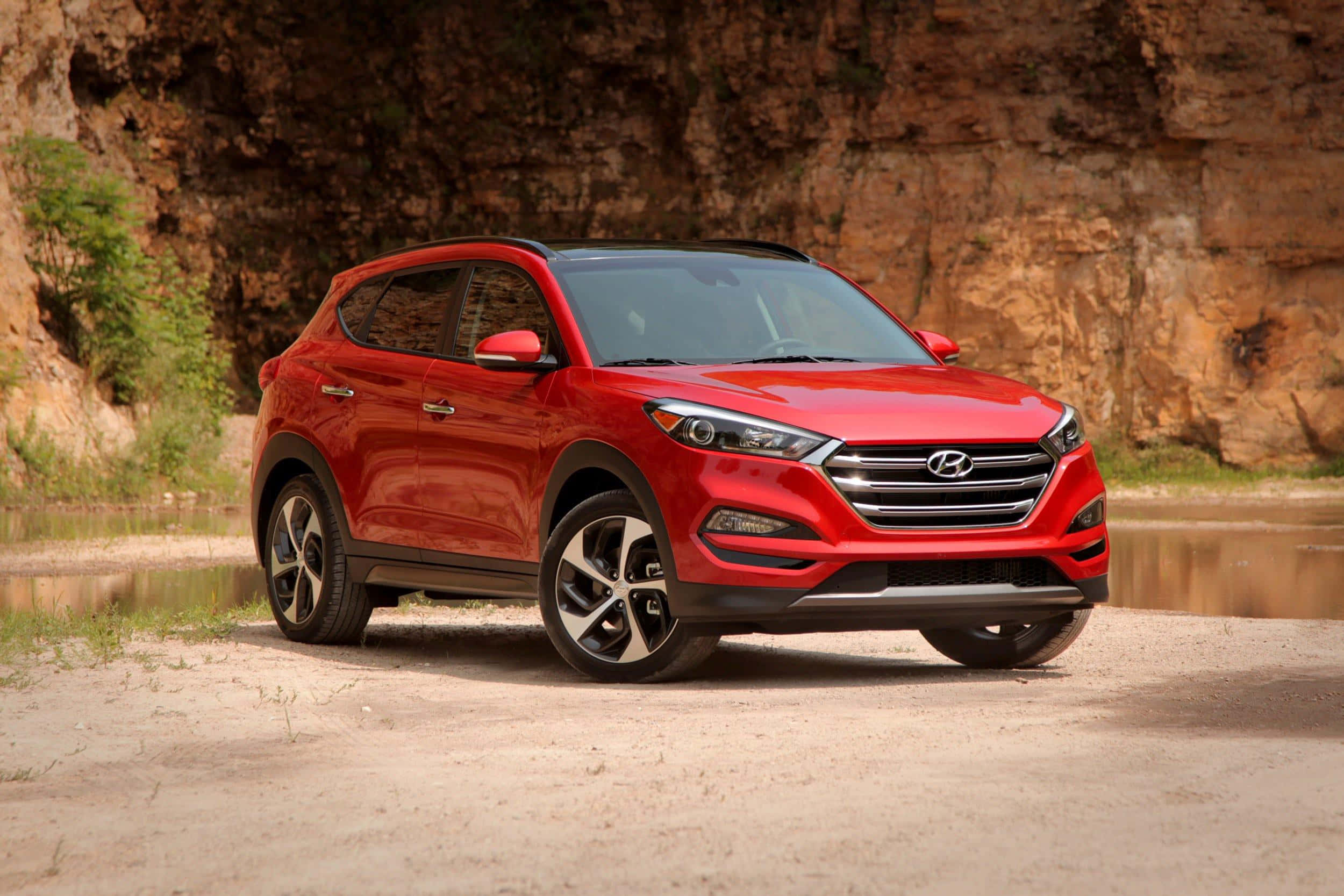 Hyundai Tucson 桌布 壁紙 — 一輛亮紅色現代Tucson SUV停泊在沙質河岸上，旁邊是岩石峭壁，綠意盎然。