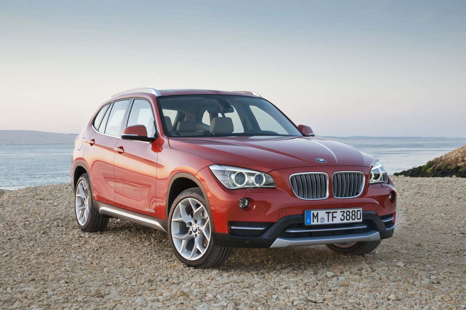 Bmw X1 桌布 壁紙 — 一輛亮紅色的BMW X1 SUV停在鵝卵石海灘上，平靜的海洋和遠山在柔和的暮色天空下。