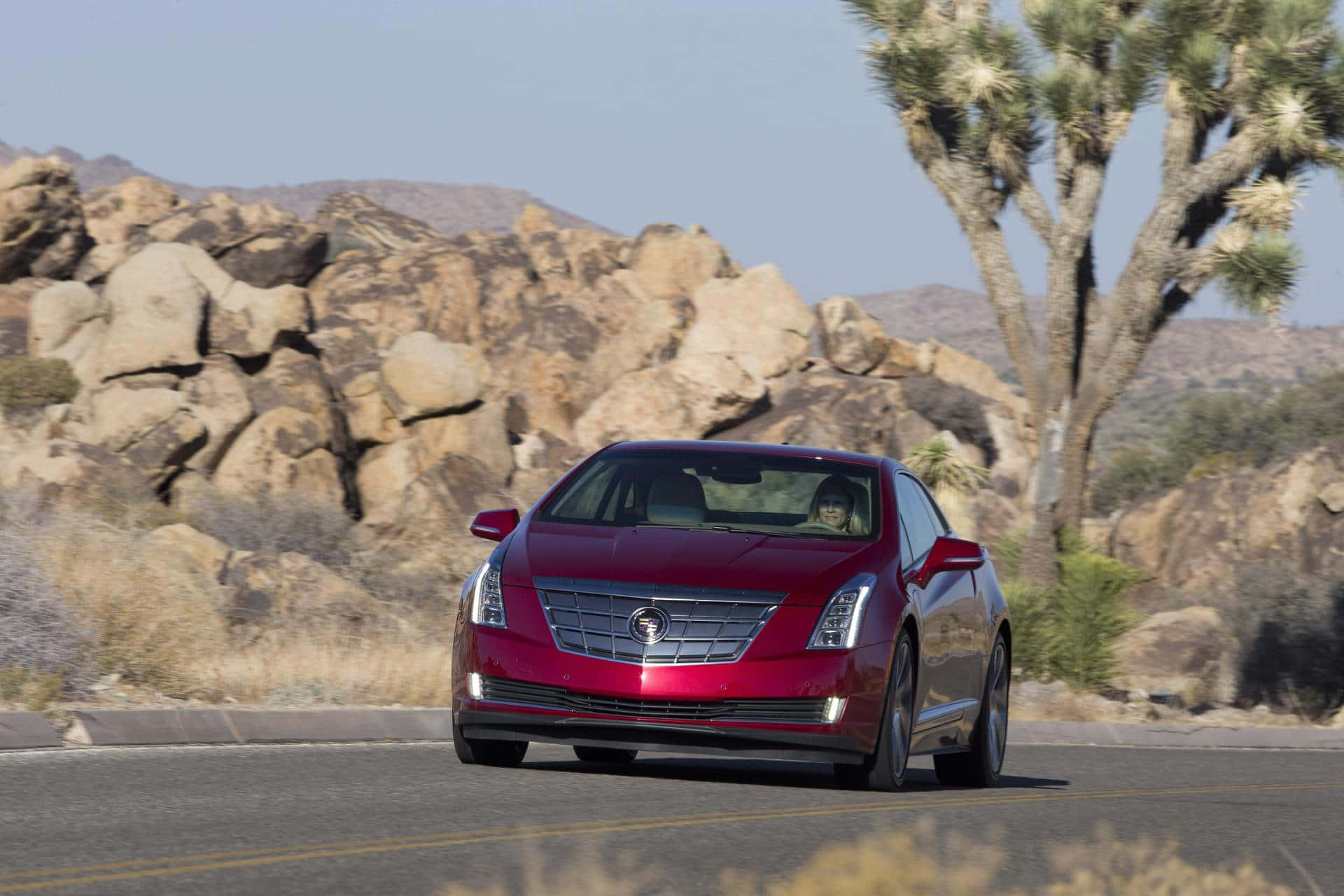 Cadillac Elr 桌布 壁紙 — 一輛亮紅色的Cadillac ELR在晴朗藍天下，沿著兩旁有岩石地形和約書亞樹的沙漠公路巡航。