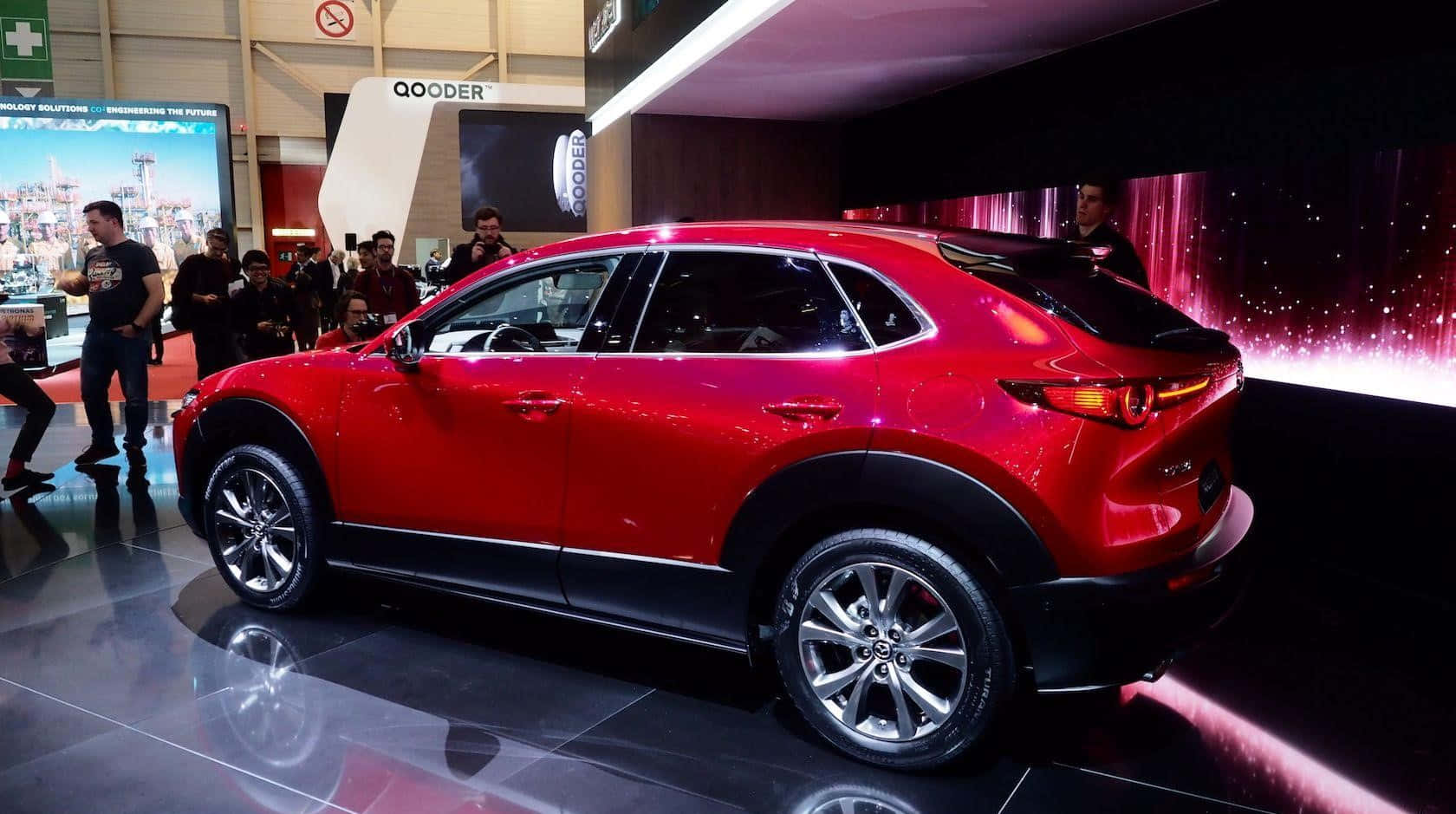 Mazda Cx-30 桌布 壁紙 — 一輛亮紅色的Mazda CX-30 SUV在繁忙的車展上，於反射地板上展示，現場燈光明亮，有參觀者。