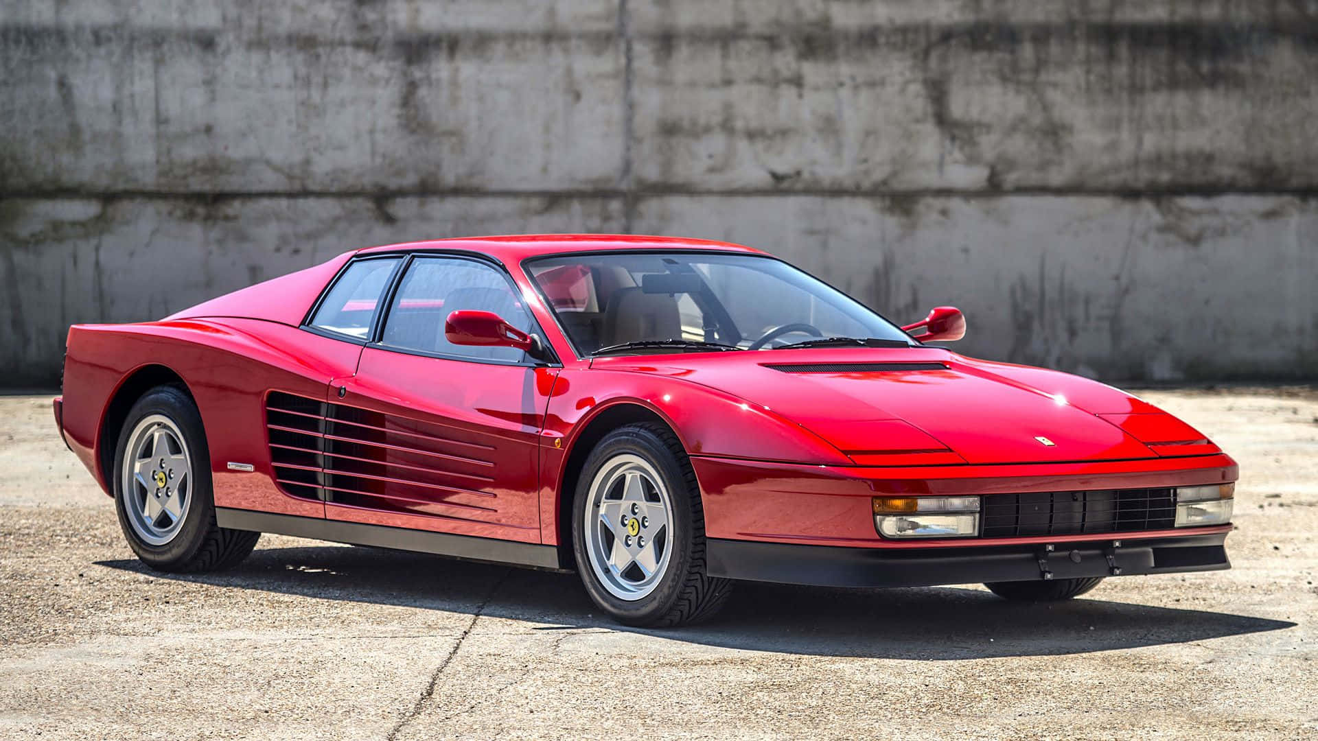 Ferrari Testarossa 桌布 壁紙 — 一輛亮紅色的法拉利 Testarossa 停泊在裂開的混凝土上，背景是風化的混凝土牆，在明亮的日光下。