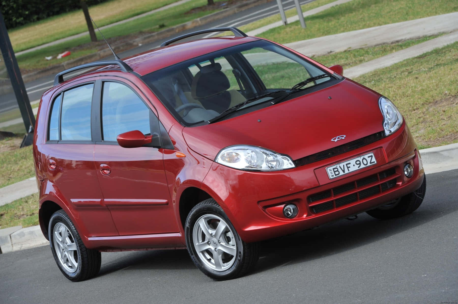Chery 桌布 壁紙 — 一輛亮紅色緊湊型汽車配有車頂行李架，停在鋪砌道路上，旁邊有草地和人行道。
