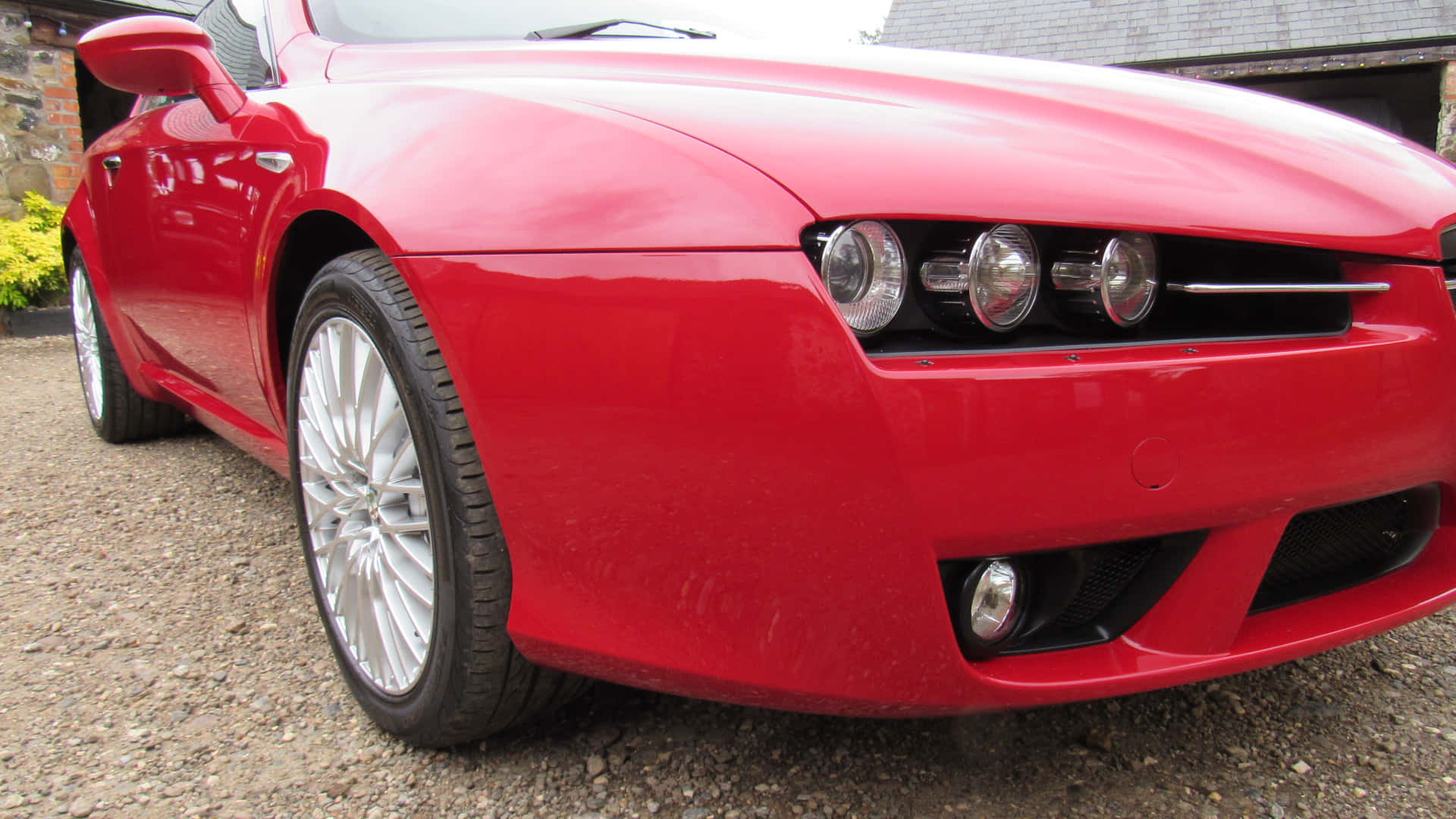 Alfa Romeo Brera 桌布 壁紙 — 一輛亮紅色跑車配銀色輪圈，停泊在石建築附近的碎石地上。