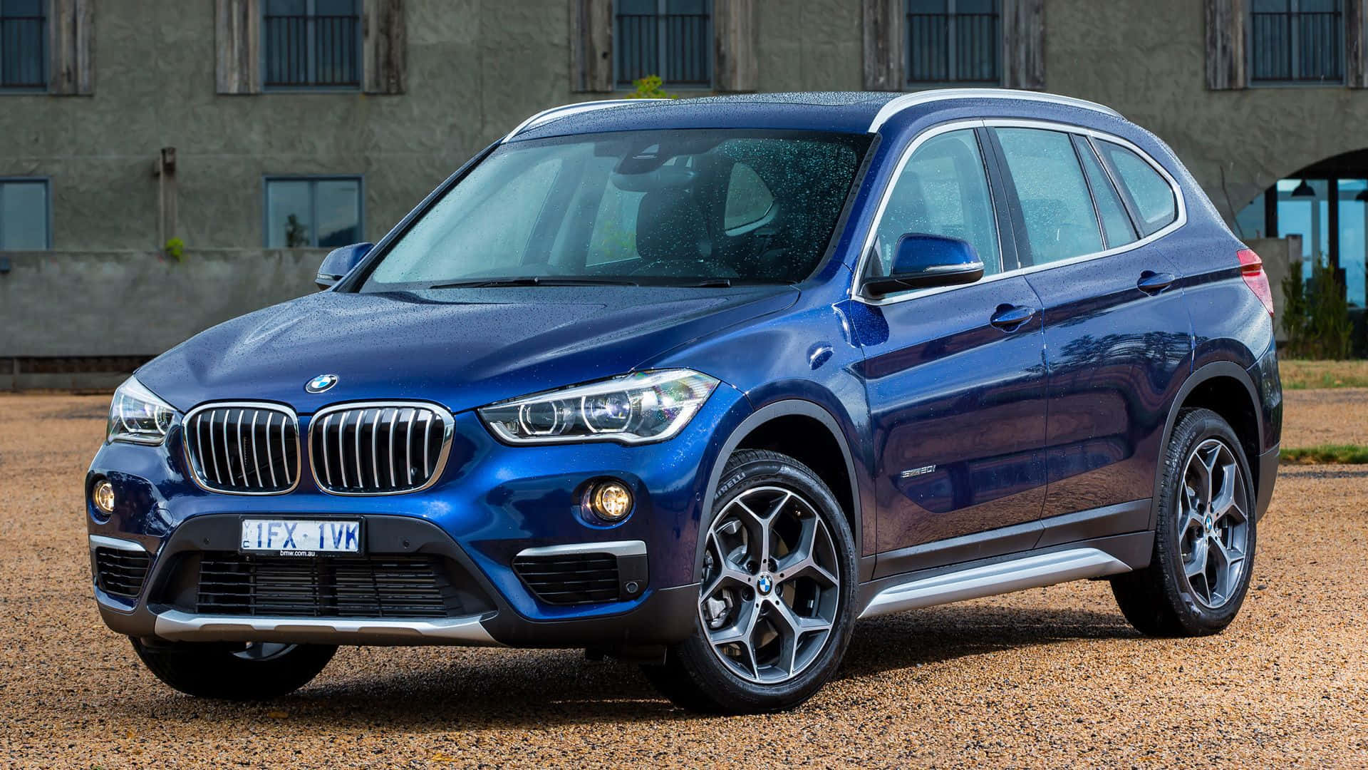 Bmw X1 桌布 壁紙 — 一輛亮藍色BMW X1 SUV停泊在風化建築物前的碎石地上，處於明亮日光下。